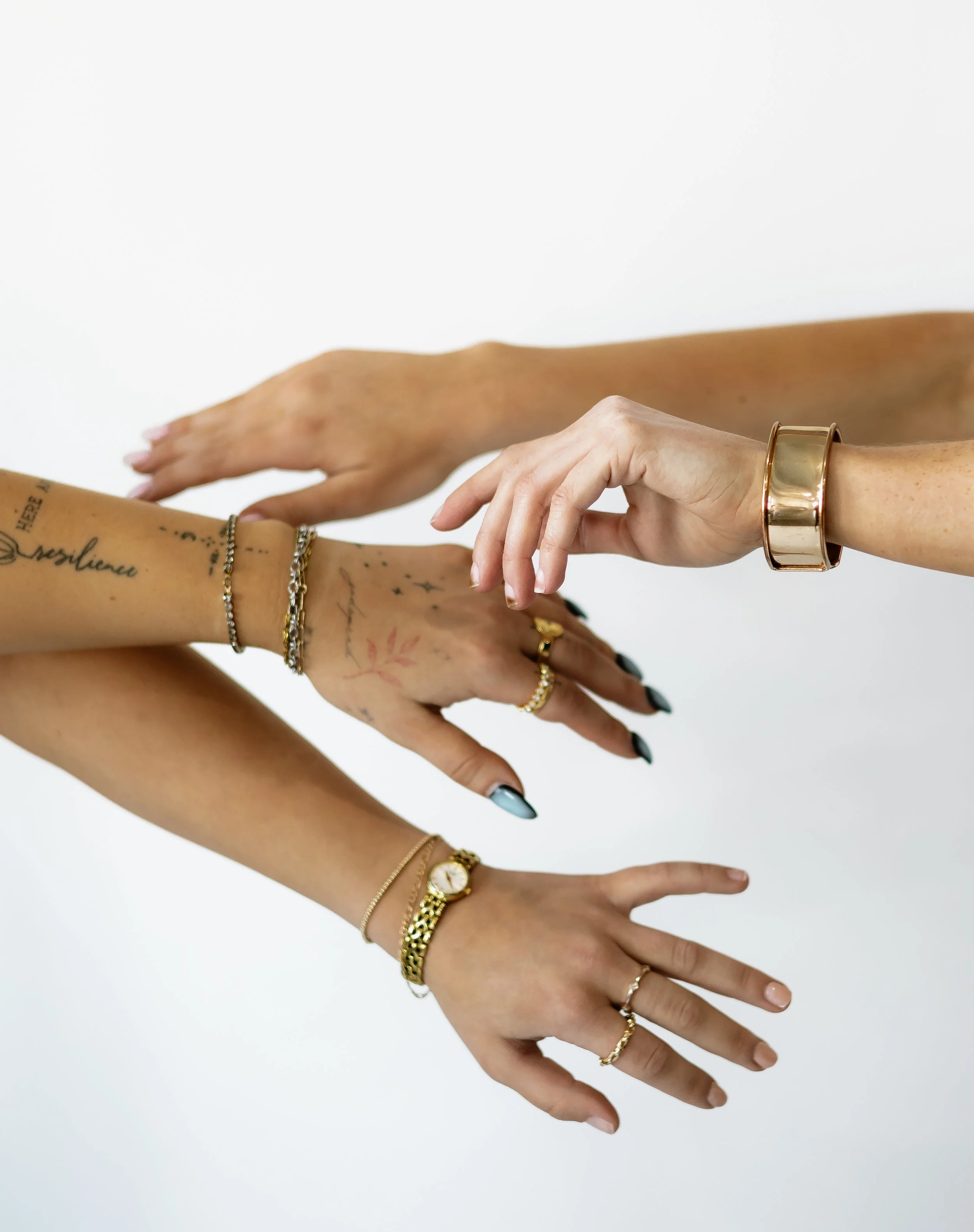 Multiple hands with jewelry and tattoos reaching towards each other against a white background.