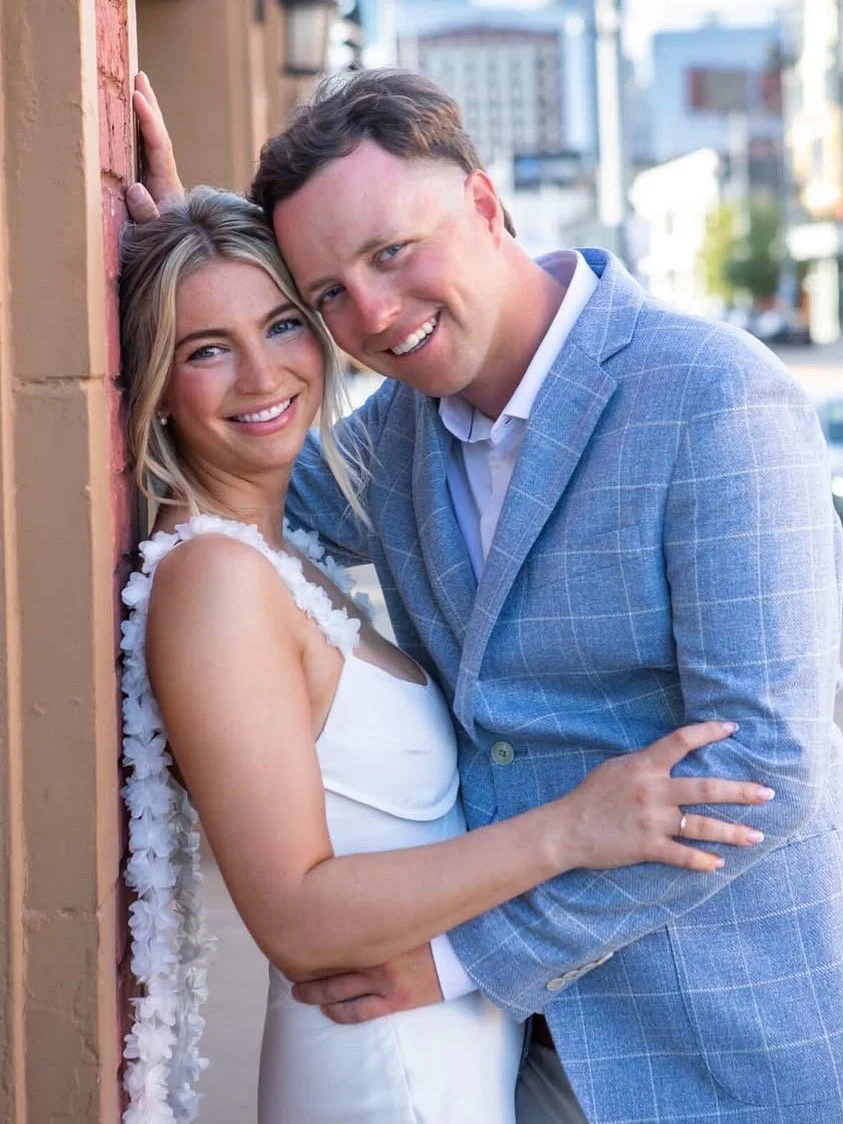 A smiling woman in a white dress with floral embellishments and a smiling man in a light blue suit, standing close together against an urban street background in daylight.