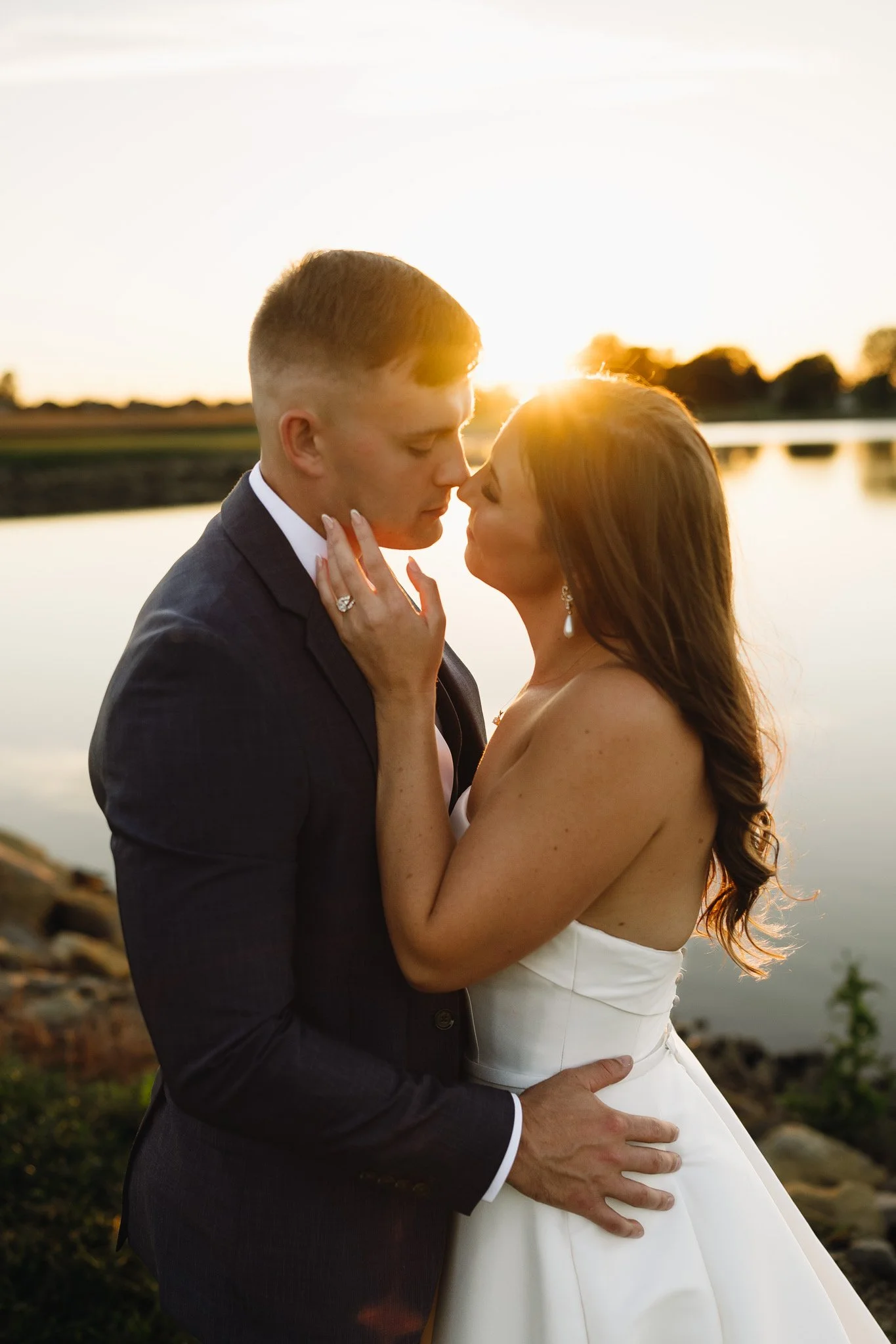 A couple at sunset, embracing by a body of water, with the woman in a white dress and the man in a dark suit, sharing an intimate moment.