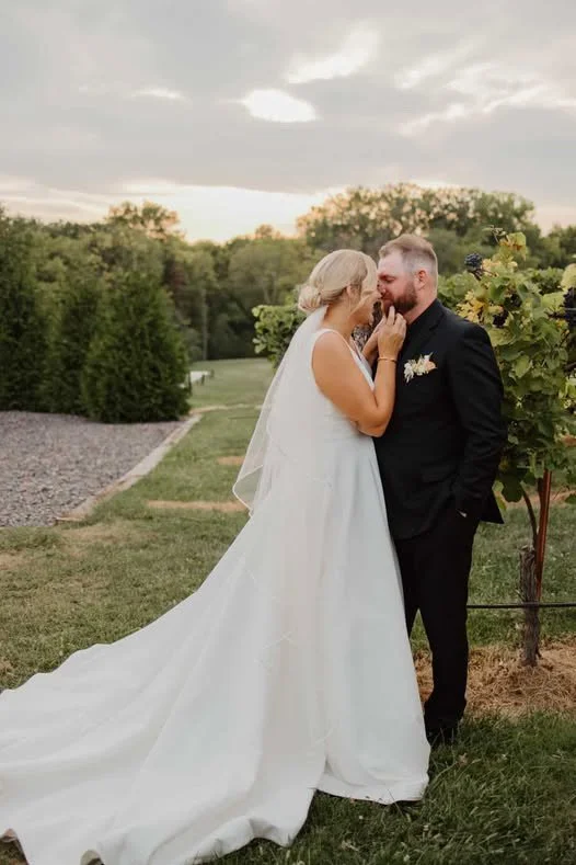 A bride and groom standing close together outdoors during sunset, in a vineyard or garden setting. The bride wears a white gown with a long veil, and the groom is in a black tuxedo with a boutonnière.