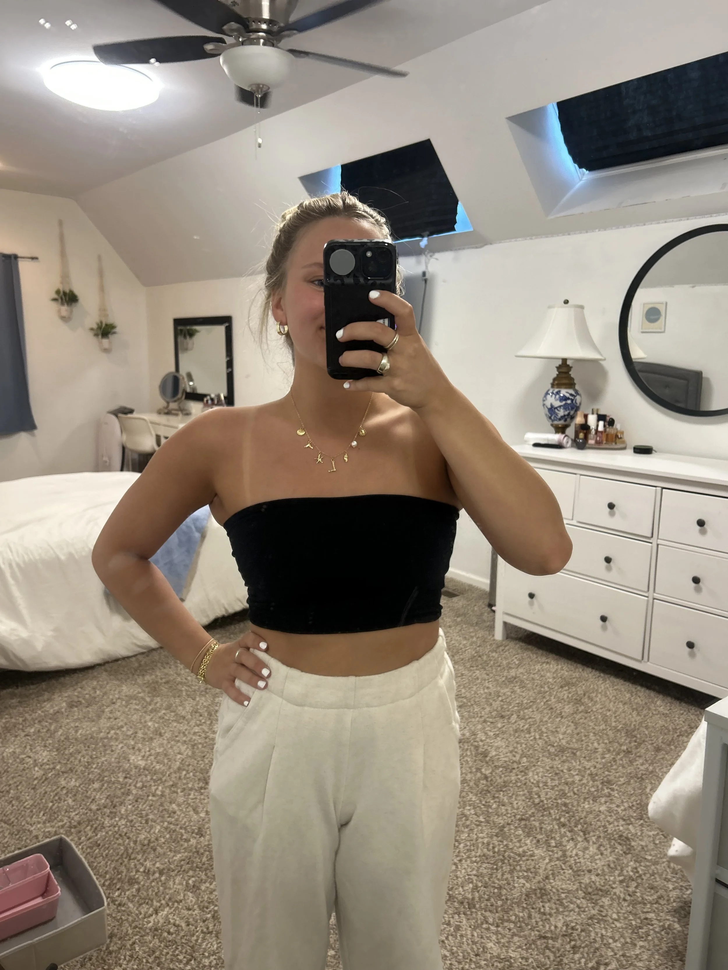A woman taking a mirror selfie in a bedroom with a sloped ceiling, ceiling fan, and attic windows. She is wearing a black tube top, cream-colored high-waisted pants, and jewelry, smiling with her hair in a messy bun.