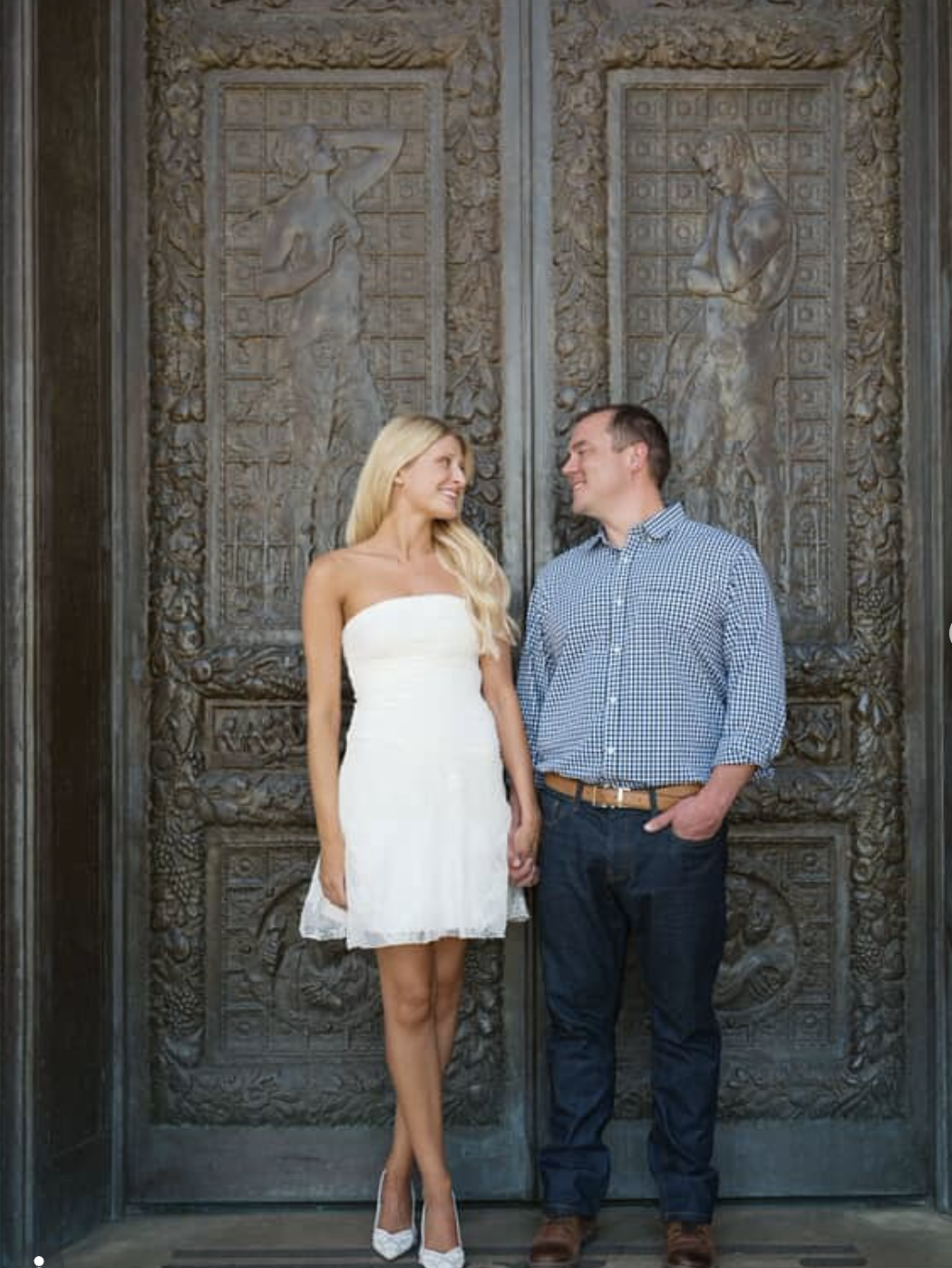 A smiling couple holding hands and looking at each other in front of ornate metal doors with intricate carvings of classical figures.