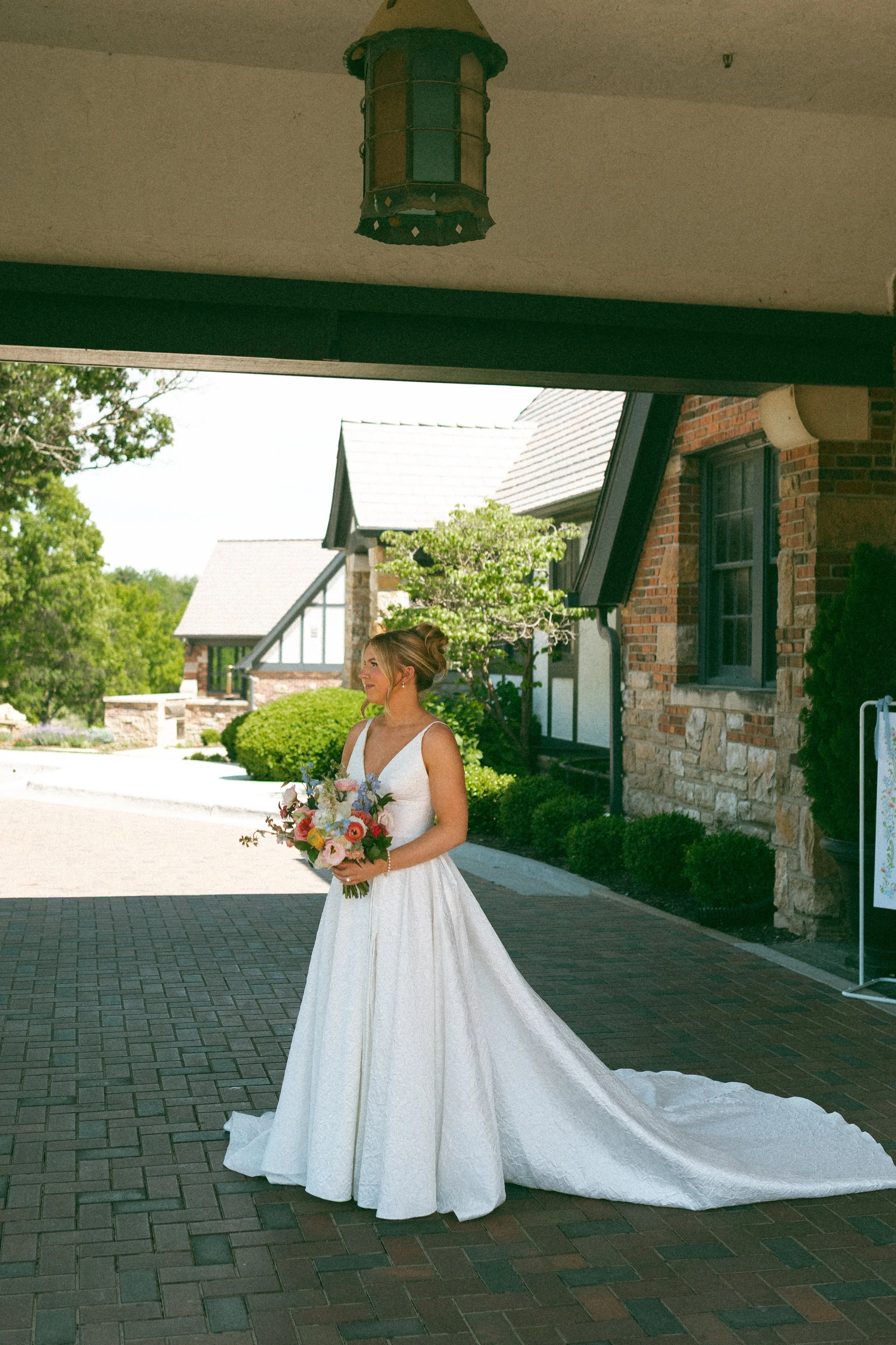 A bride in a white wedding dress holding a colorful bouquet standing outside under a covered area, with a brick building and green trees in the background.