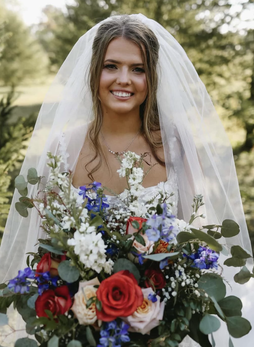 A smiling bride in a white wedding dress with a veil, holding a large colorful bouquet of flowers, outdoors with trees in the background.