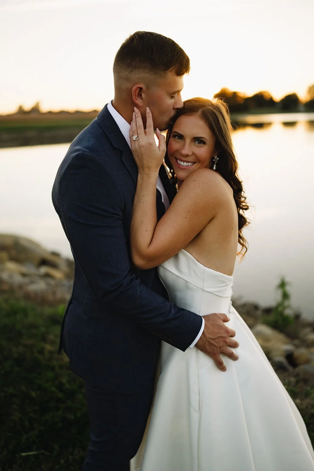 A newlywed couple sharing a romantic moment outdoors by a lake during sunset, with the man in a dark suit and the woman in a white strapless wedding dress, smiling and embracing each other.