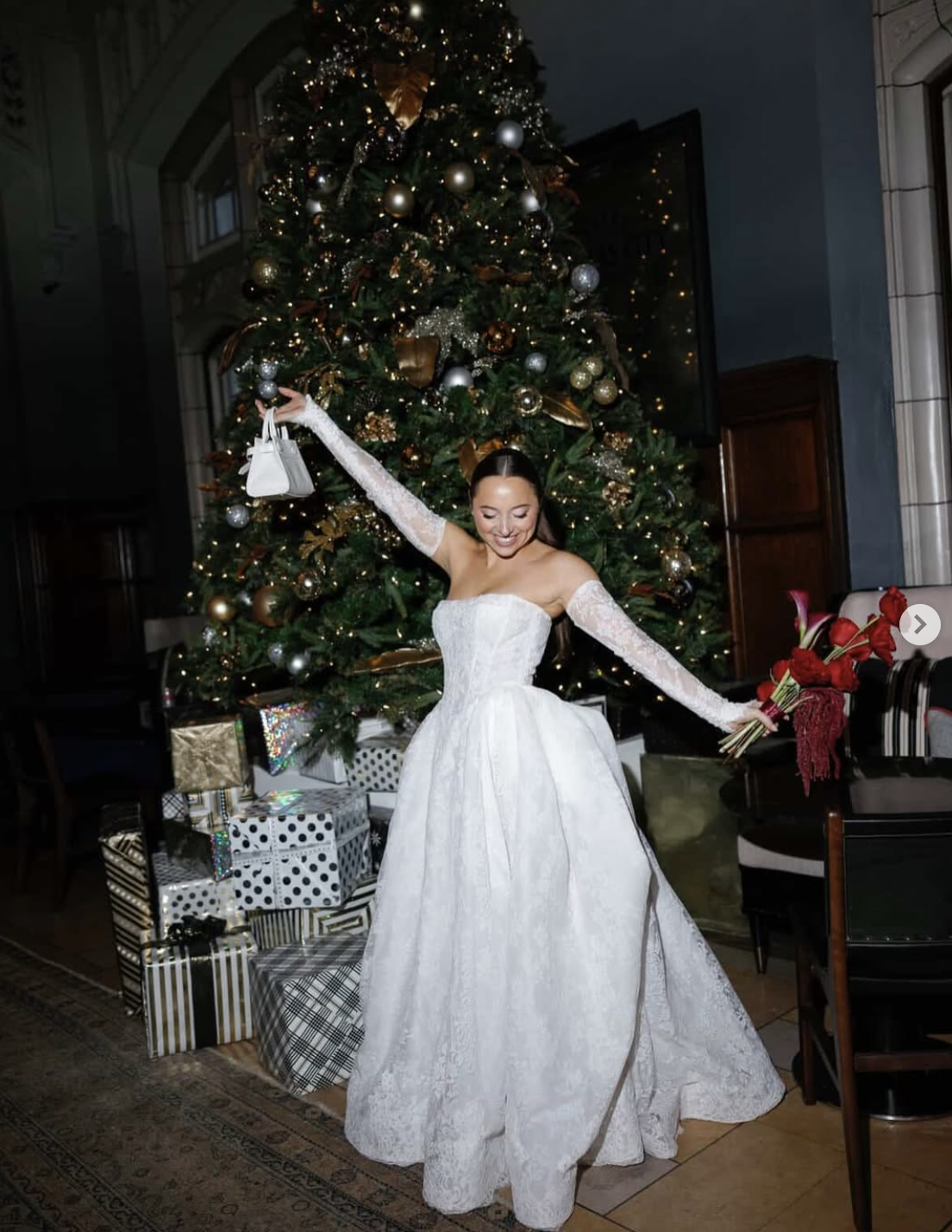 A woman in a white strapless wedding gown with lace gloves stands joyfully in front of a decorated Christmas tree holding a bouquet of red flowers in one hand and a small white purse in the other. There are wrapped presents underneath the tree.