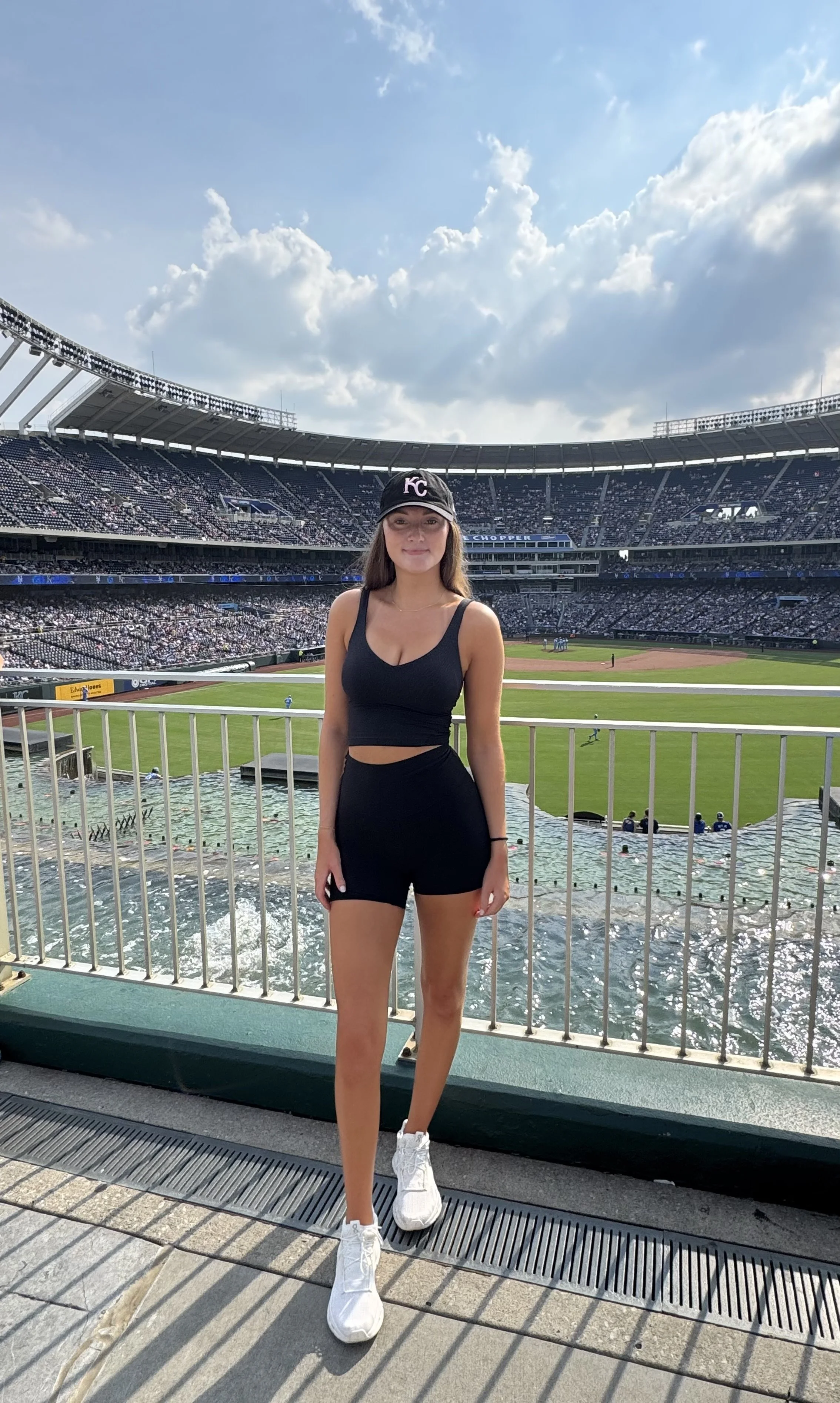 Young woman in black sportswear and a Kansas City baseball cap standing in front of a stadium with a baseball field, crowd, and an outfield waterfall feature.