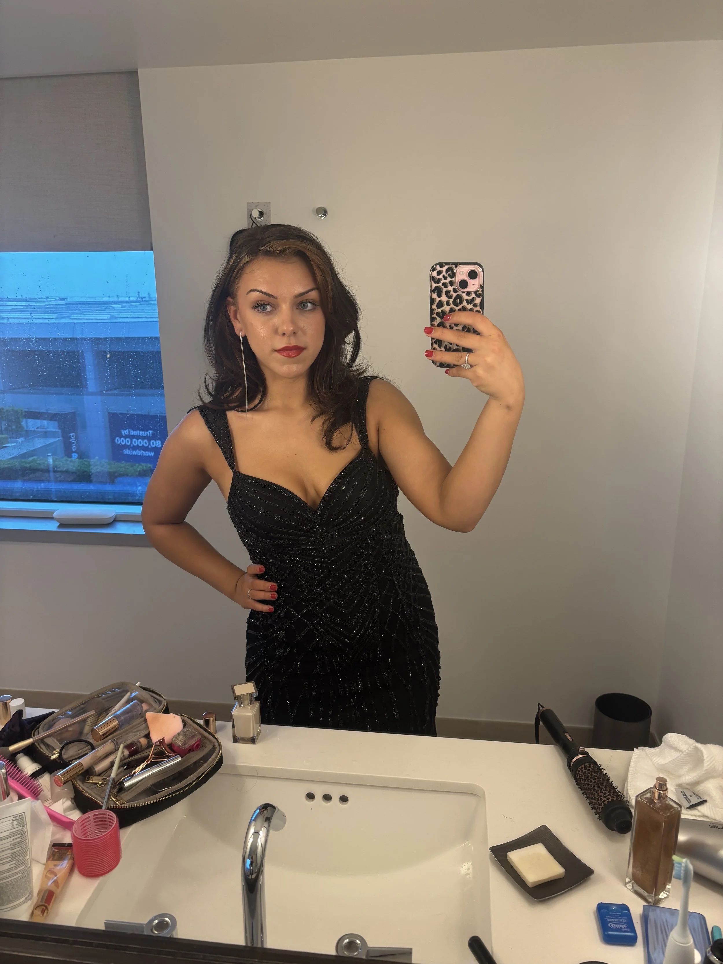 A woman taking a selfie in a bathroom mirror, wearing a black dress with spaghetti straps.