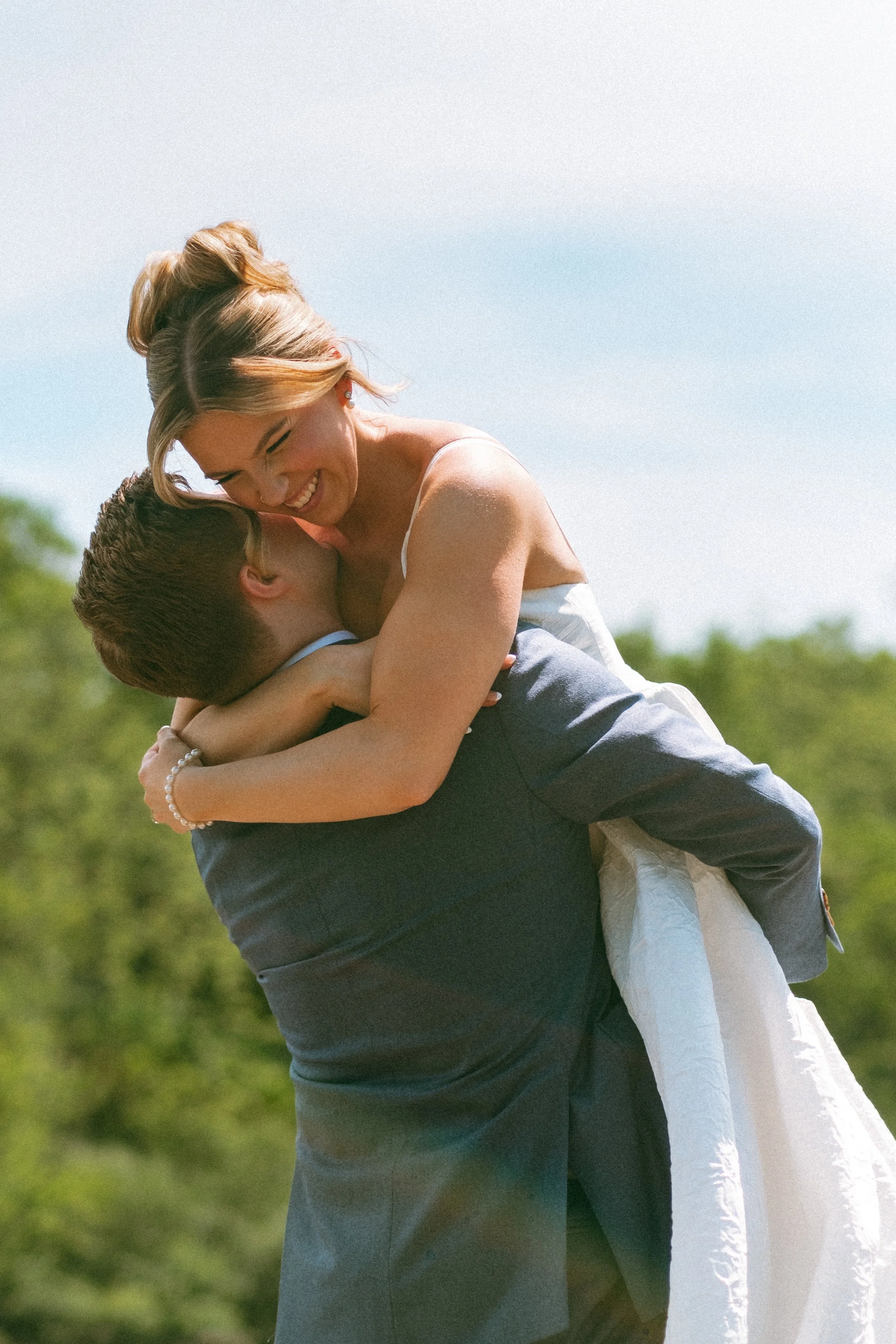 A woman in a wedding dress is being lifted by a man in a suit, both smiling and enjoying a joyful moment outdoors with trees in the background.