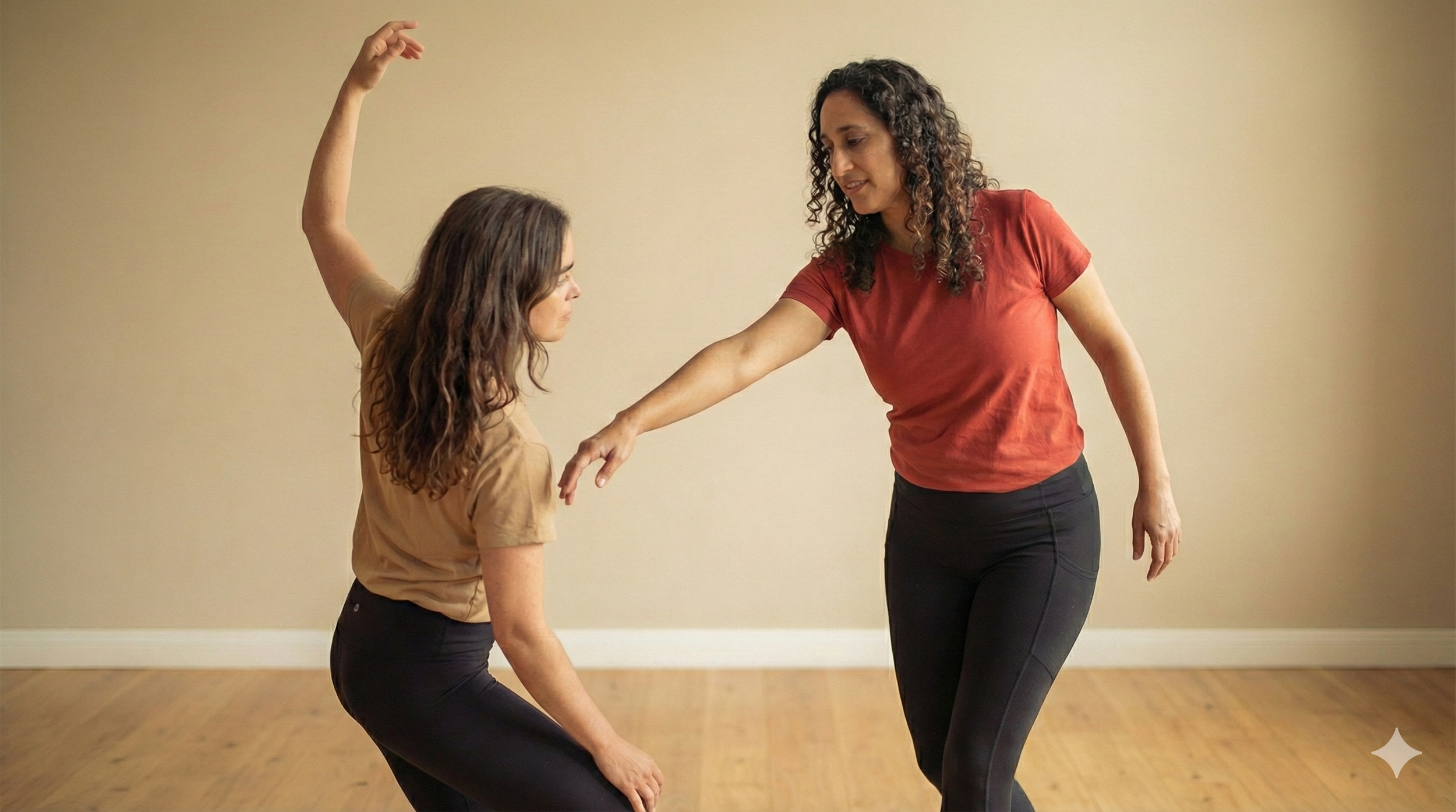A woman in a red shirt is guiding a young woman in a brown shirt through a dance or movement exercise in a room with wooden floor and beige walls.