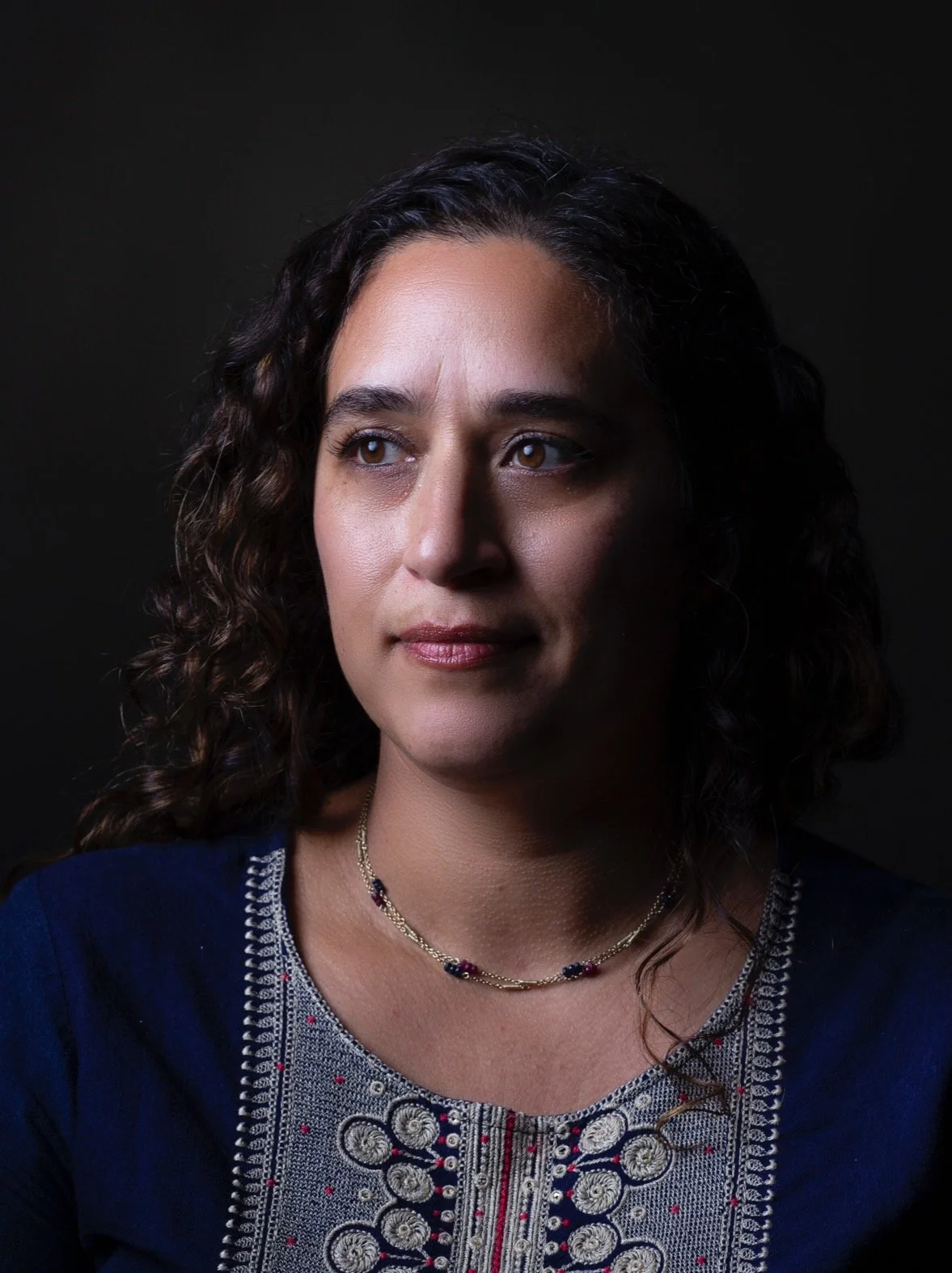 A woman with curly dark hair wearing a blue embroidered top and a necklace posed against a dark background.