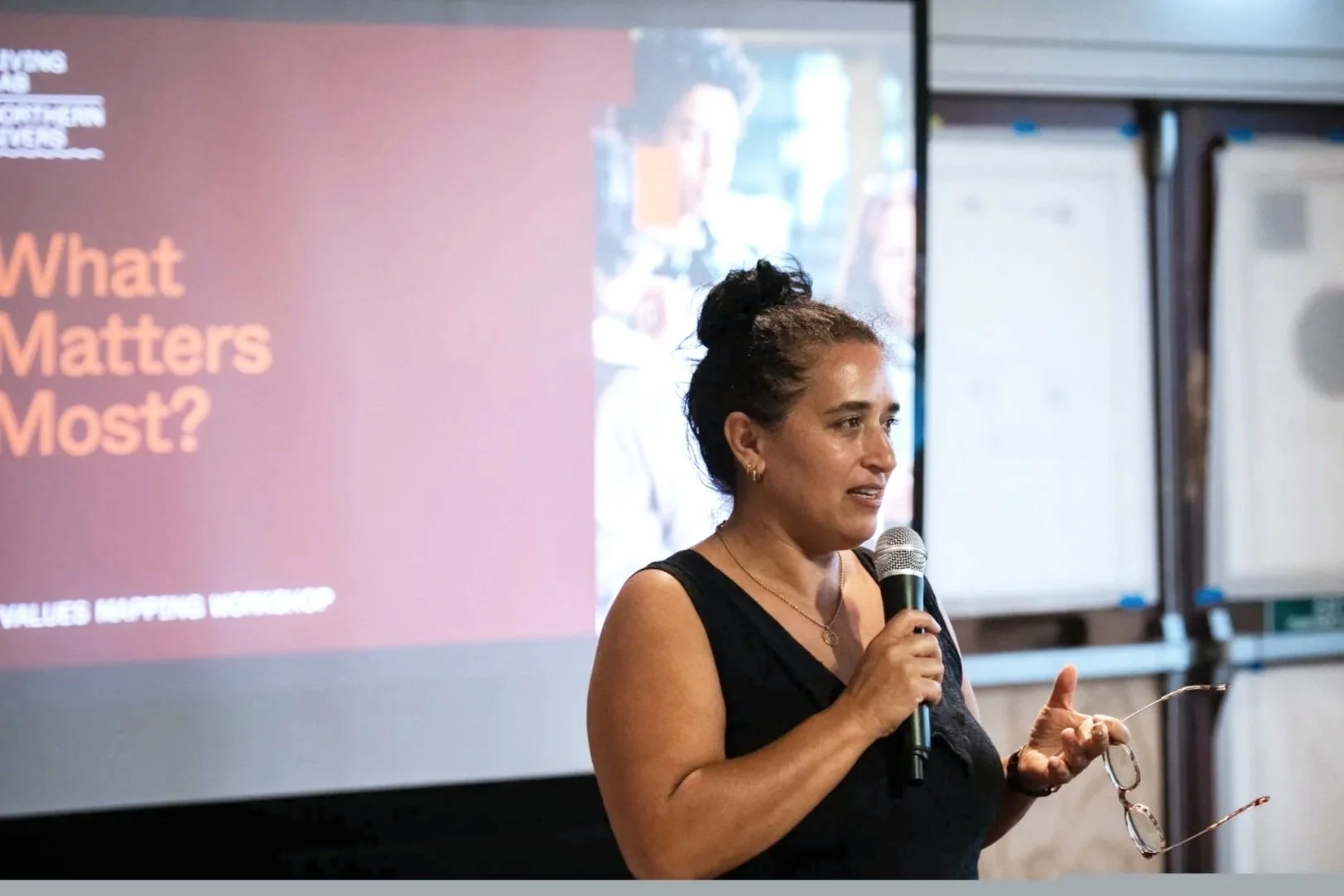 A woman in a black sleeveless top speaking into a microphone during a presentation, with a slide titled 'What Matters Most?' displayed on a projector screen behind her.