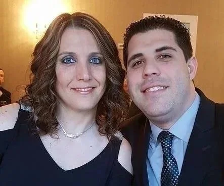 A woman with shoulder-length curly hair and blue eyes smiling, wearing a black dress and pearl necklace, standing next to a man with short dark hair, wearing a light blue dress shirt, dark suit, and polka dot tie, both in an indoor setting with a warm background.