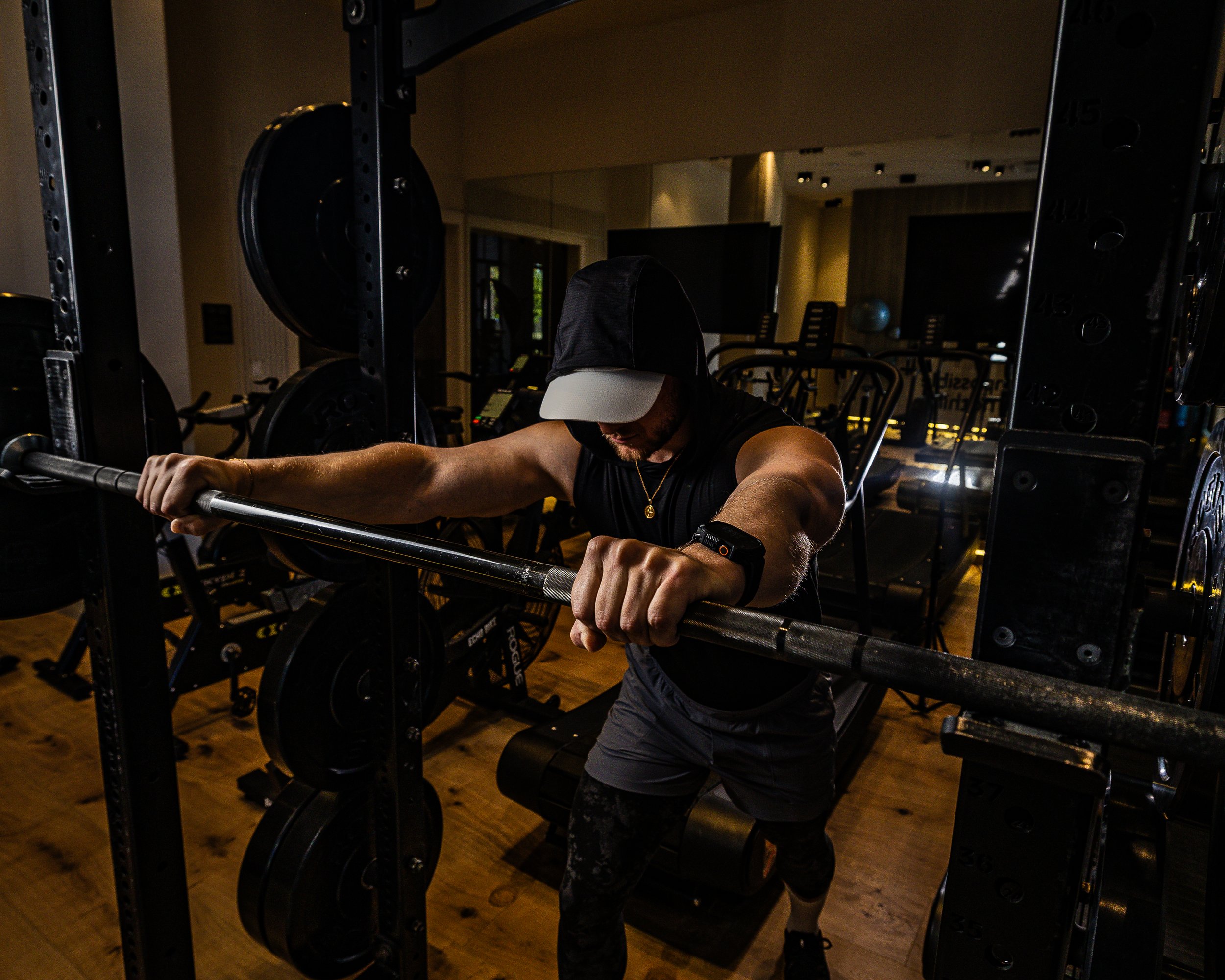 A man wearing a black hoodie, white cap, and black workout clothes lifting a barbell in a gym.
