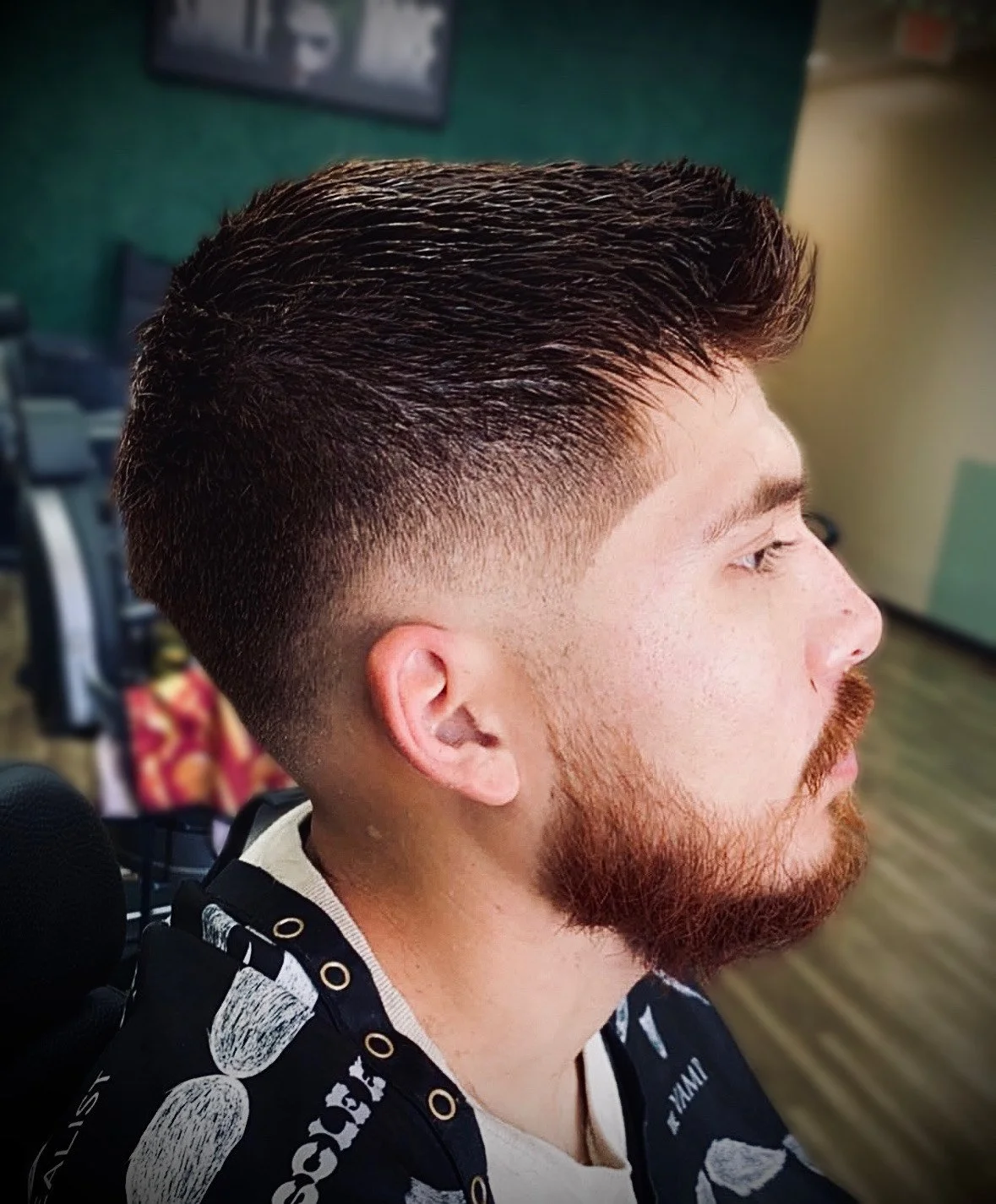 Side profile of a young man with a short textured haircut and a reddish-brown beard, sitting in a barber shop.