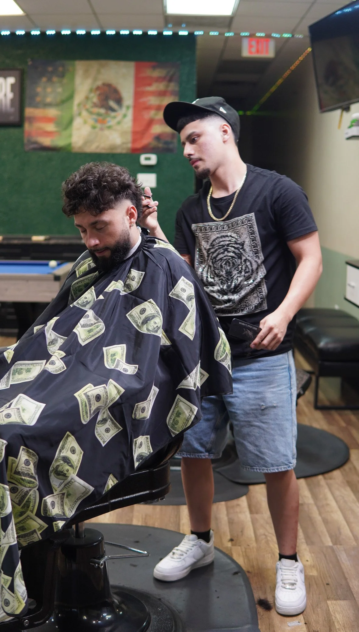 A man with a beard is getting a haircut in a barber shop, covered with a cape with dollar bill patterns. A barber wearing a black T-shirt with a tiger design, denim shorts, and a cap is cutting his hair. There is a Mexican flag on the wall in the background.