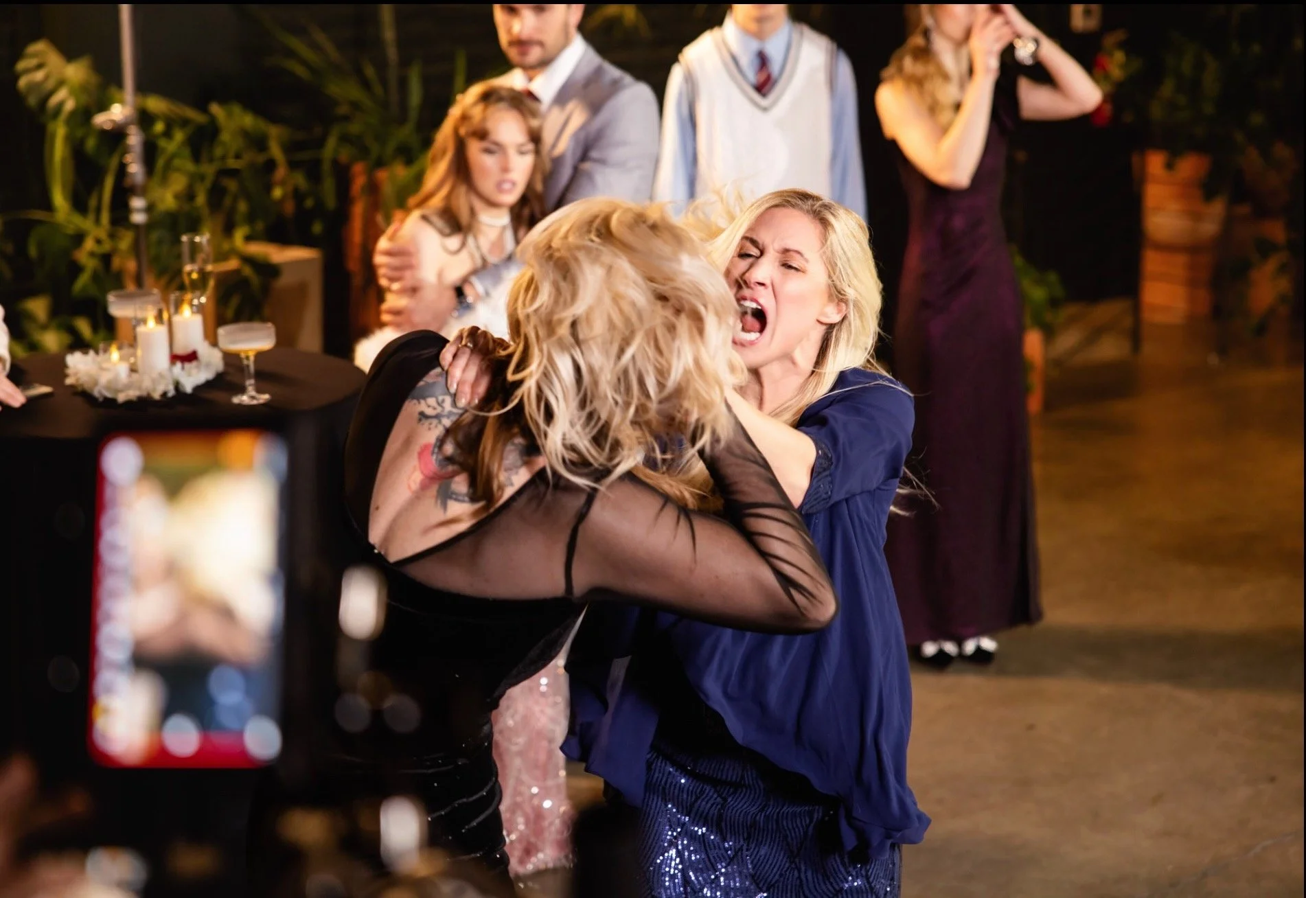 Two women fighting passionately at a party, with one woman grabbing the other by the shoulders and the other woman showing a loud, angry expression. In the background, several people are watching, including a man in a gray suit and a woman on a phone in a dark purple dress, with candles and decorative plants on nearby tables.