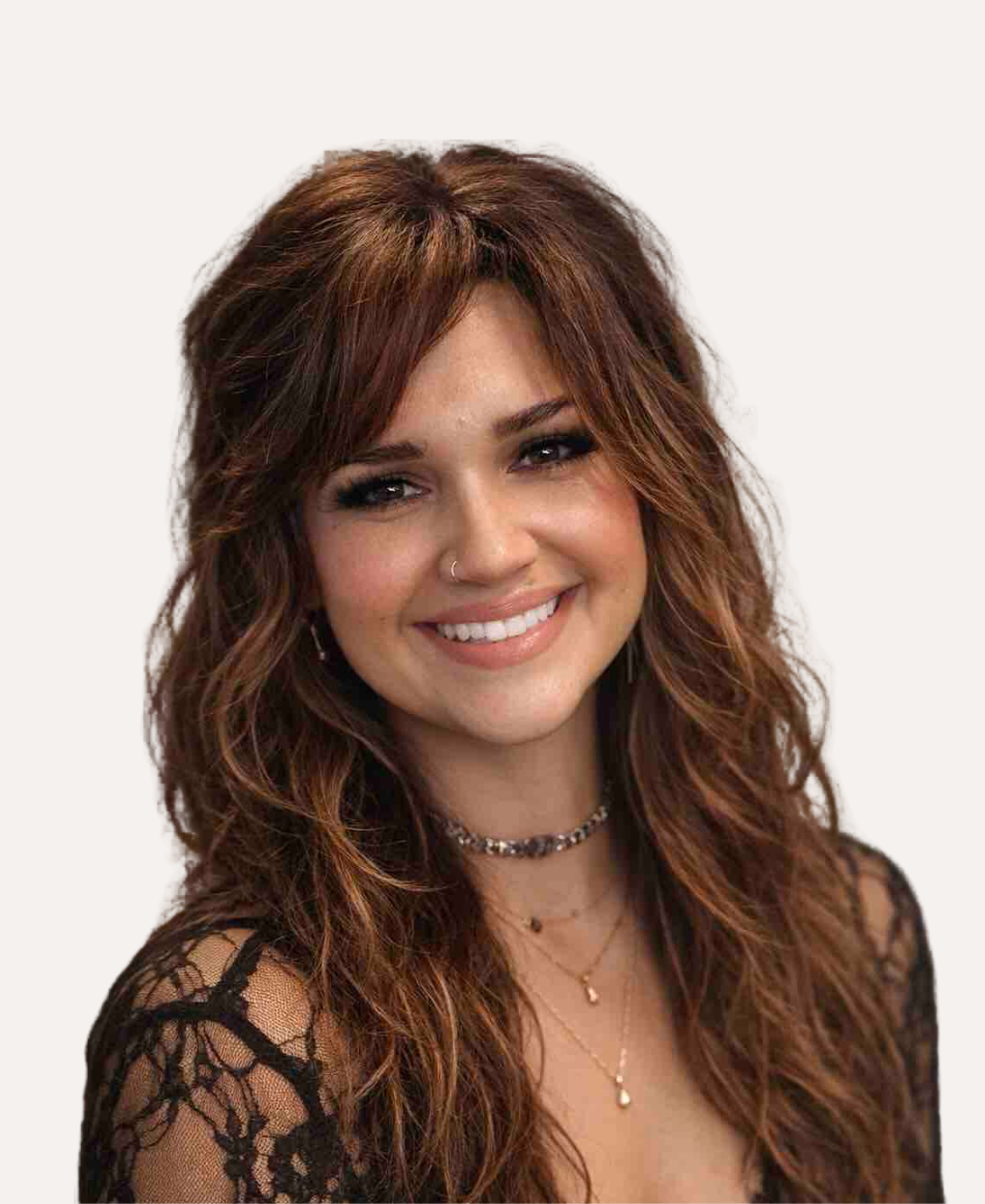 A woman smiling with long brown wavy hair, wearing a black top, standing against a plain off-white background.