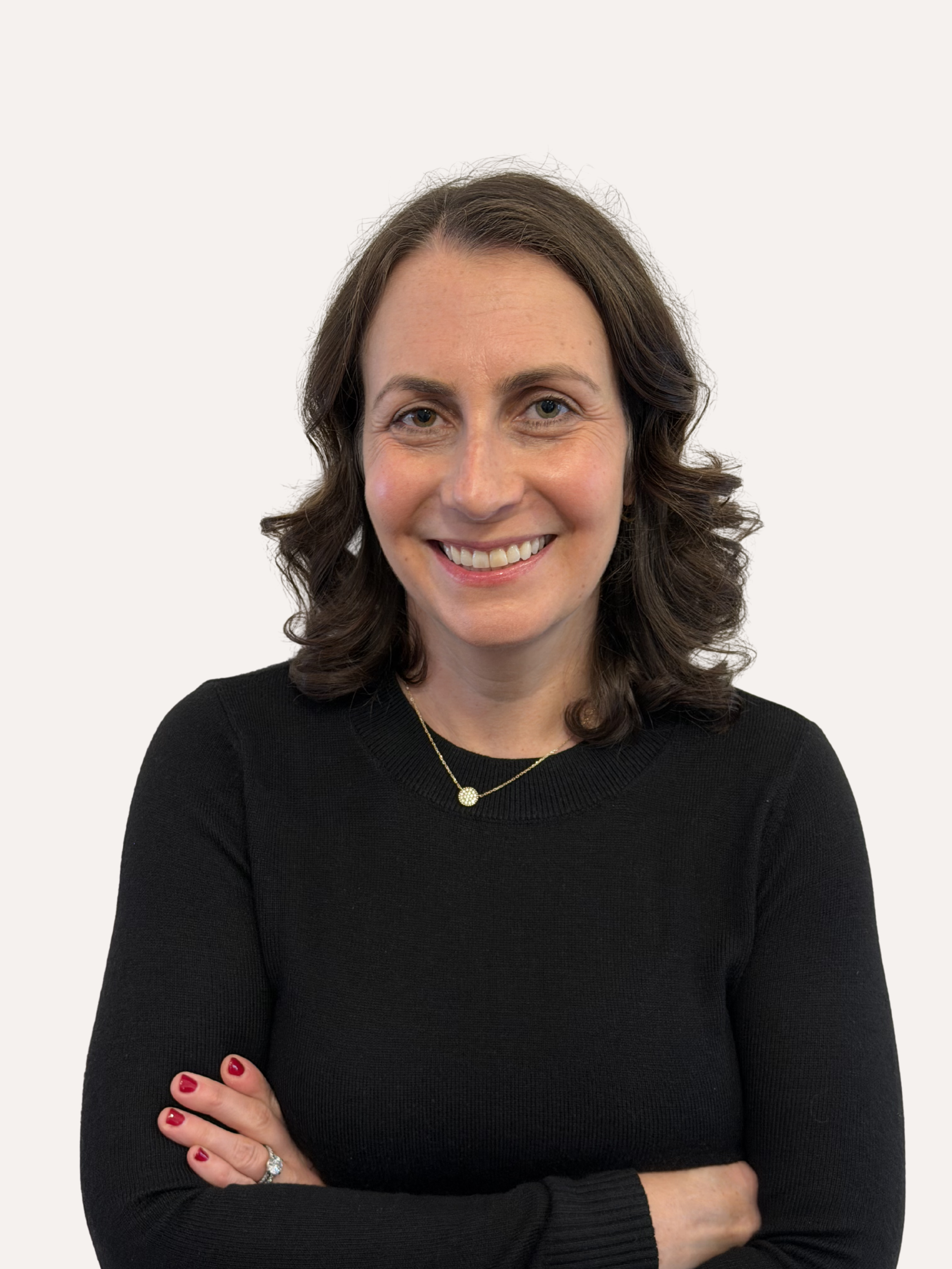 A woman with dark wavy hair, and gold necklace wearing a black textured sweater, smiling with arms crossed against a plain light background.