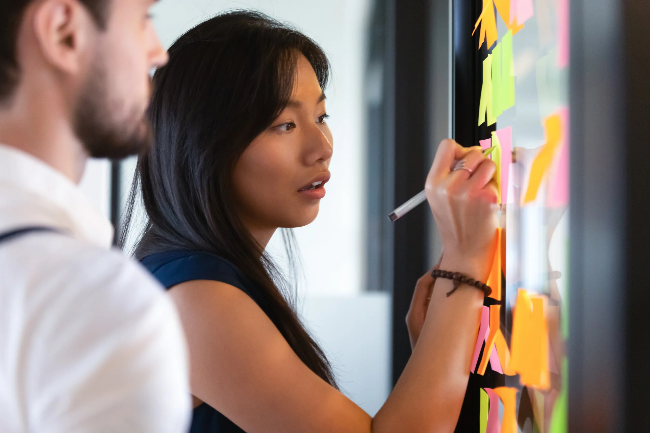 Sticky notes on a window with a woman writing on one of them while a man standing next to her observes, presumably her team member