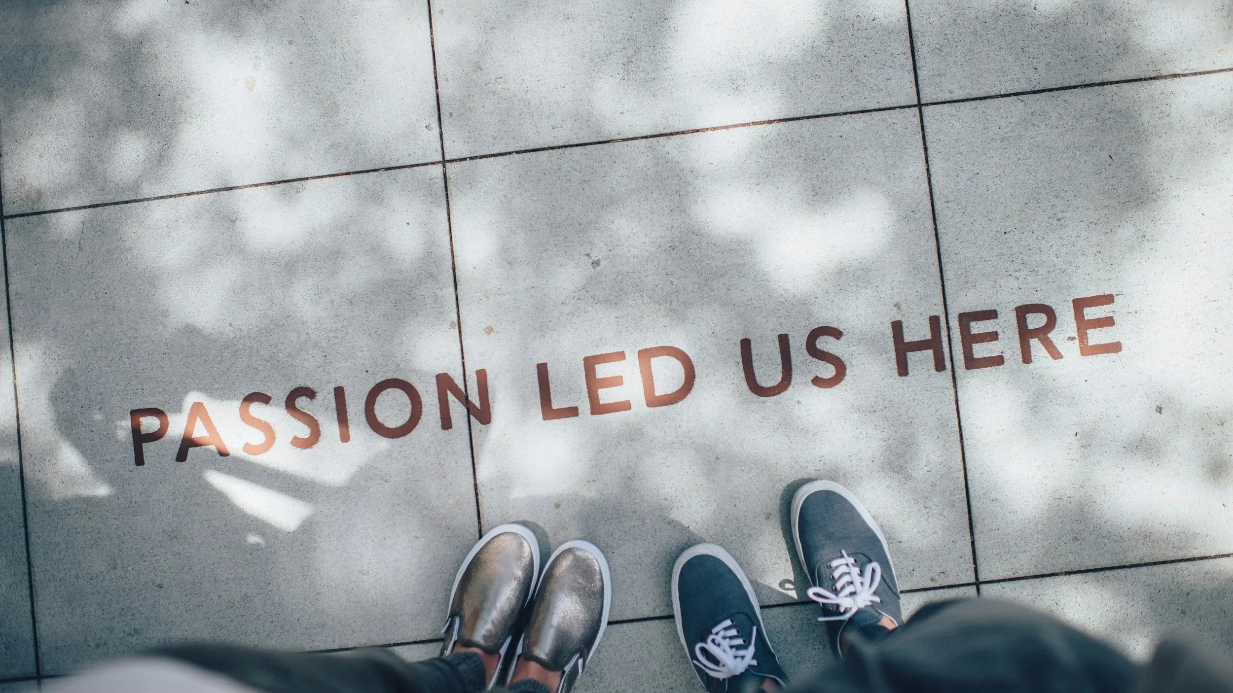 The photo shows a tiled sidewalk with shadows cast by tree leaves, and the words 'PASSION LED US HERE' written in orange above the shoes of two people standing on the sidewalk.