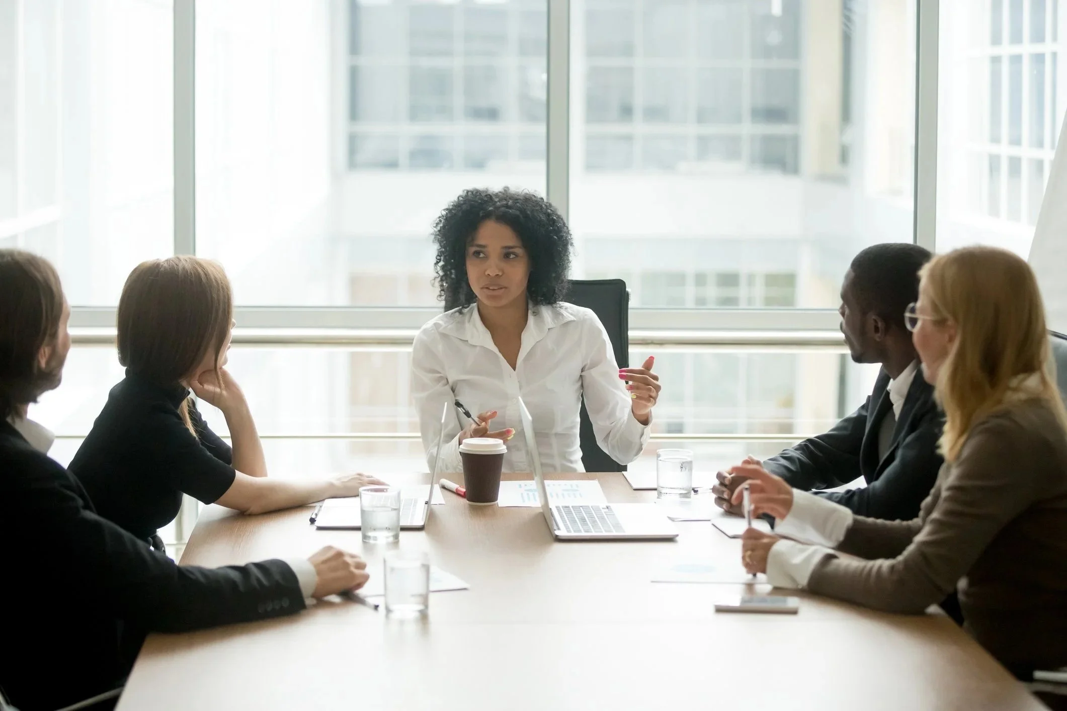 Team of five at a conference table with one person talking and gesturing and others listening.