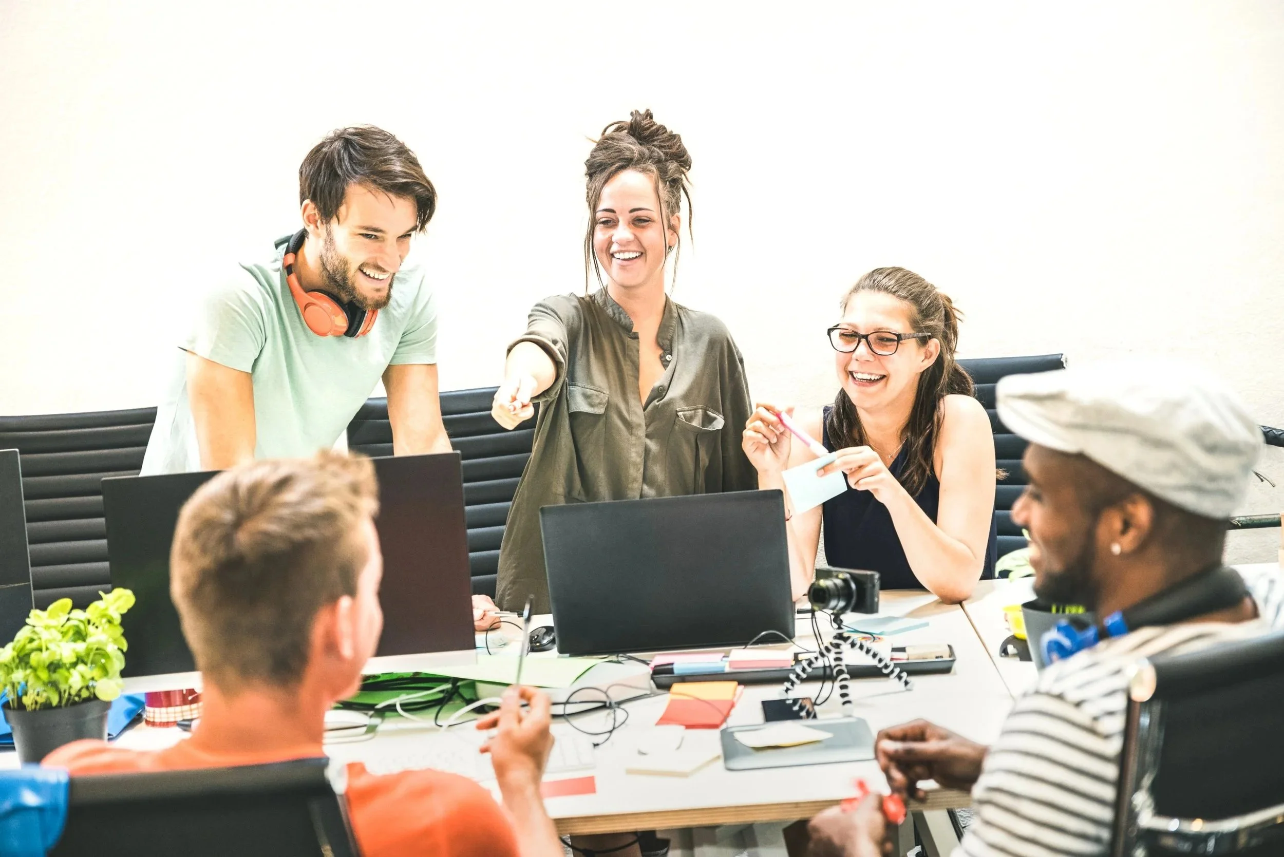 A group of people gathered around a table in a meeting, people smiling and engaging, with laptops, notebooks, and plants on the table, in a bright, modern room.