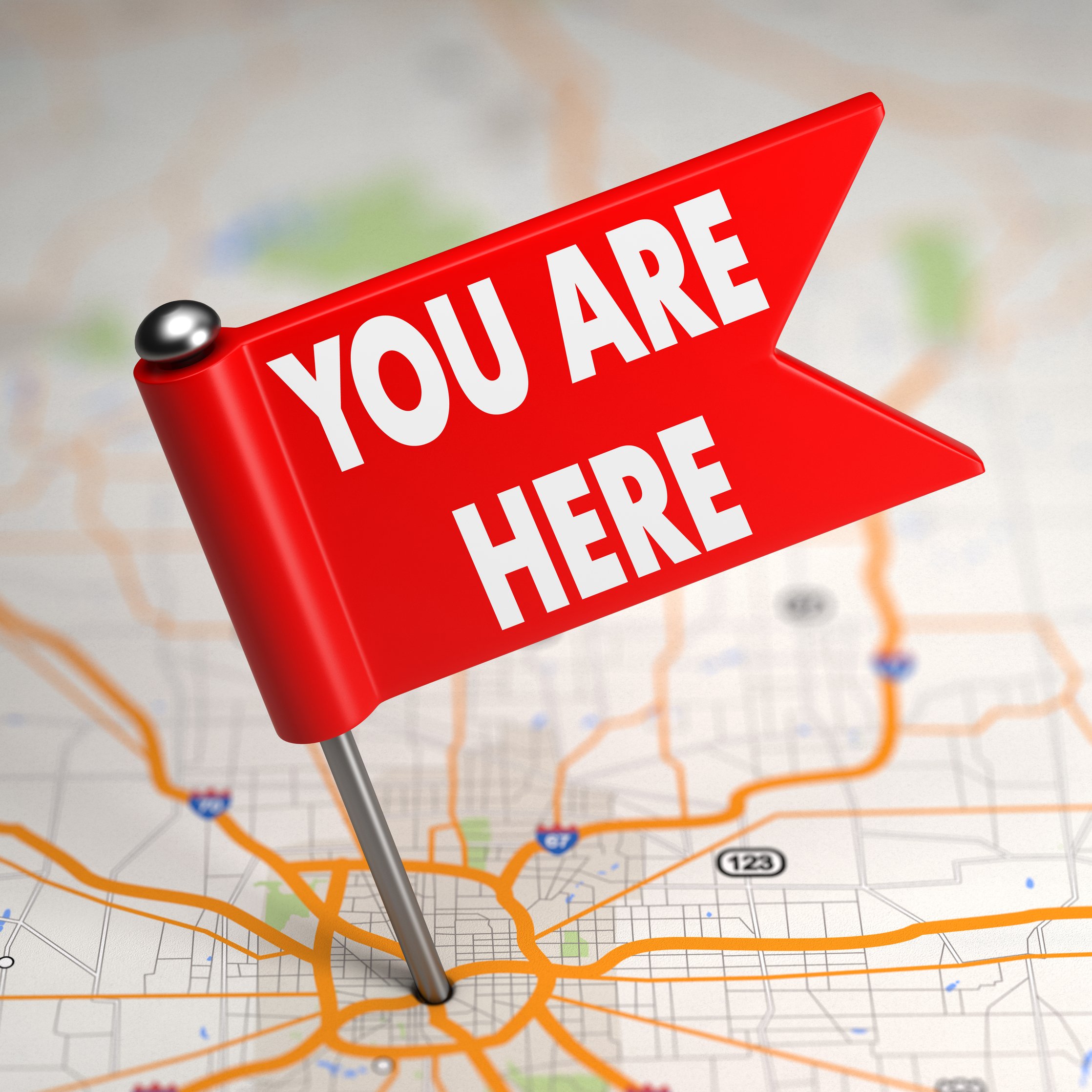 A red sign shape with the words 'You Are Here' in white, positioned on a map of a city with orange and black street lines and green areas in the background.