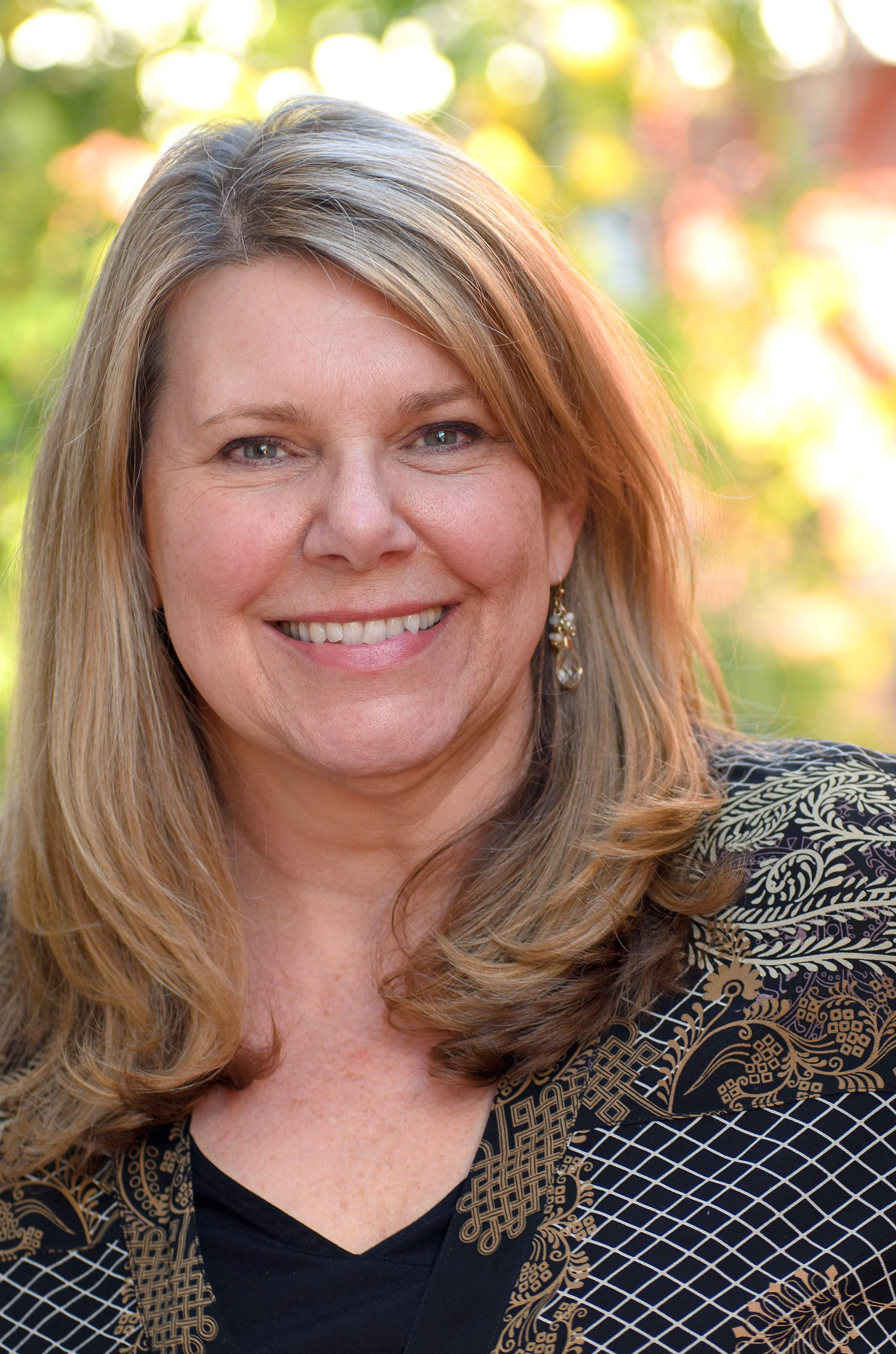 A smiling middle-aged woman with long, wavy blonde hair and green eyes, wearing a black top and a black and gold patterned jacket, standing outdoors with blurred greenery and sunlight in the background.