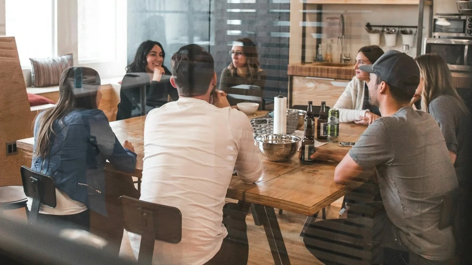 A diverse group of seven people, a team, sitting around a wooden conference table in a modern conference room, having a conversation.