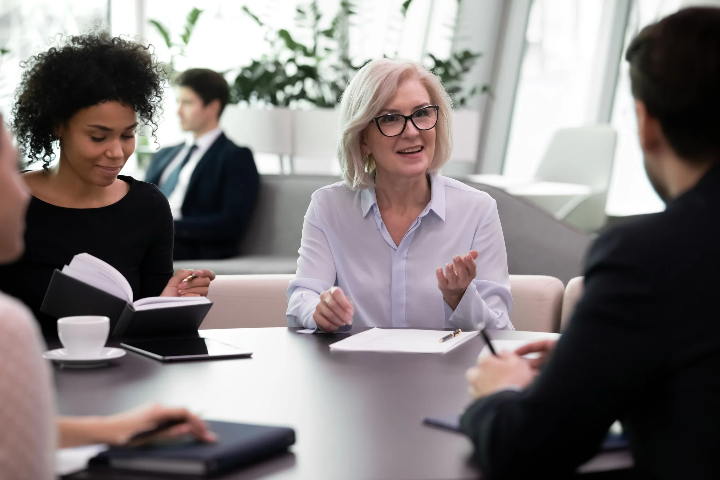 Four people sitting at a round conference table, one woman making notes, another woman in glasses speaking and smiling, and a woman and a man listening.