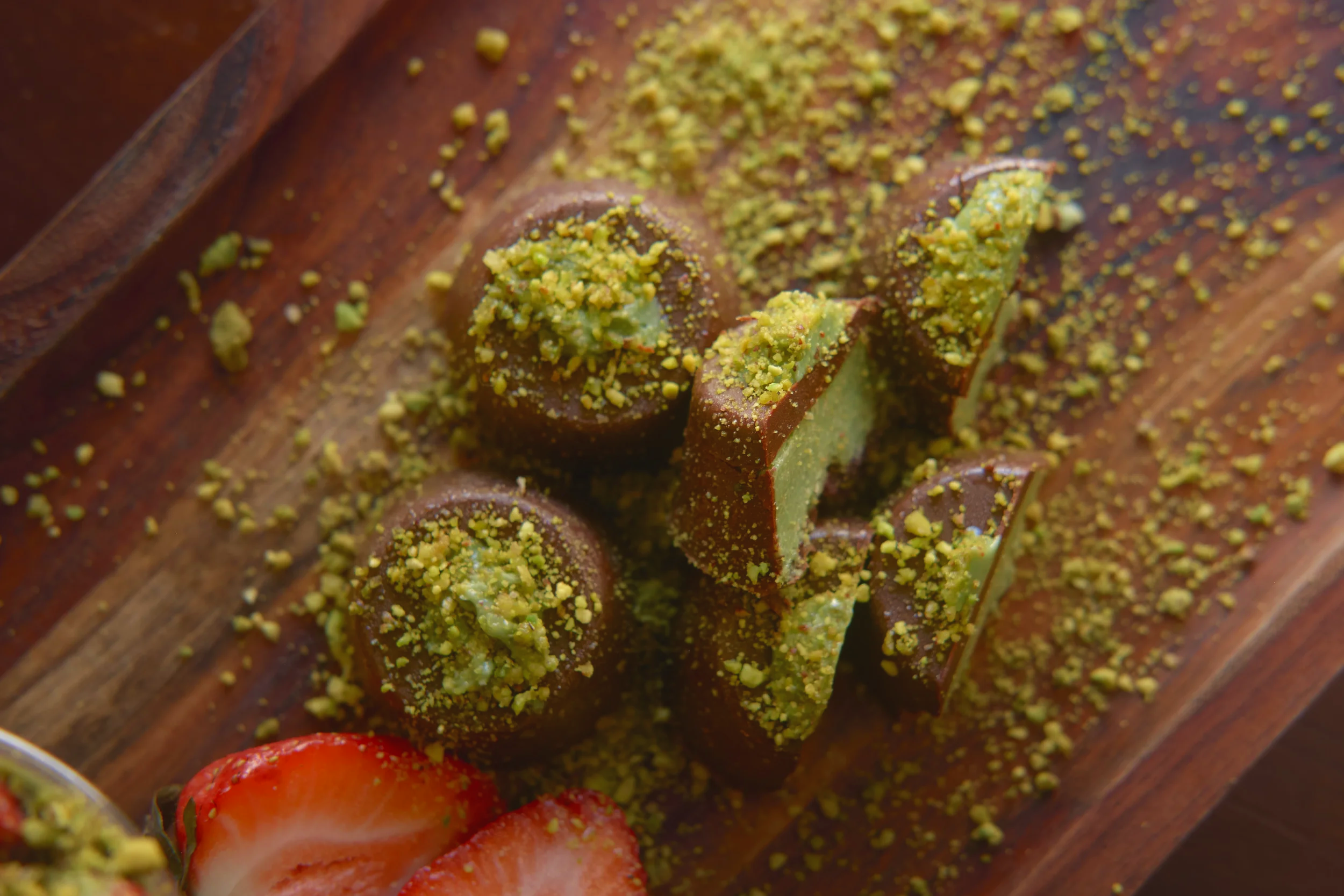 Close-up of a chocolate dessert topped with finely chopped pistachions, with sliced strawberries and cherry tomato in the foreground.