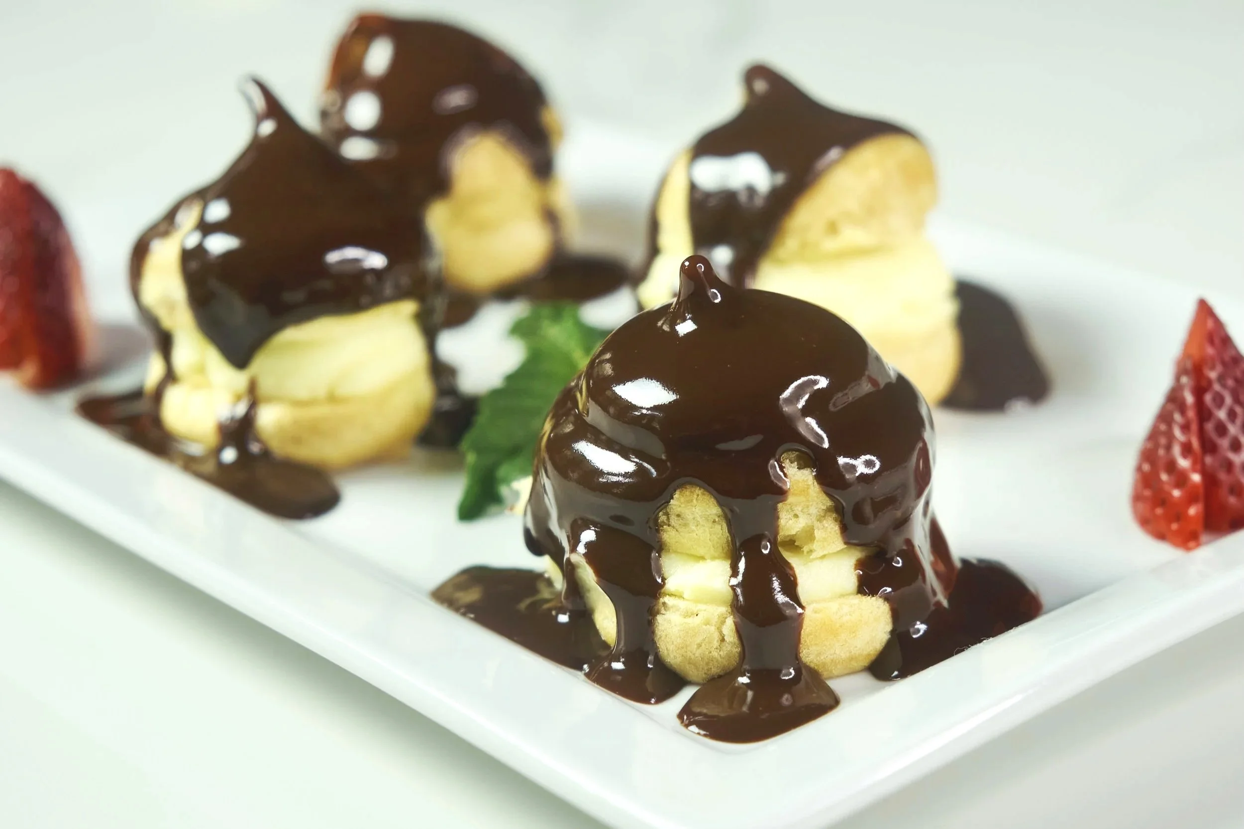 Cream puffs topped with chocolate sauce, garnished with strawberries and fresh mint, served on a white platter.
