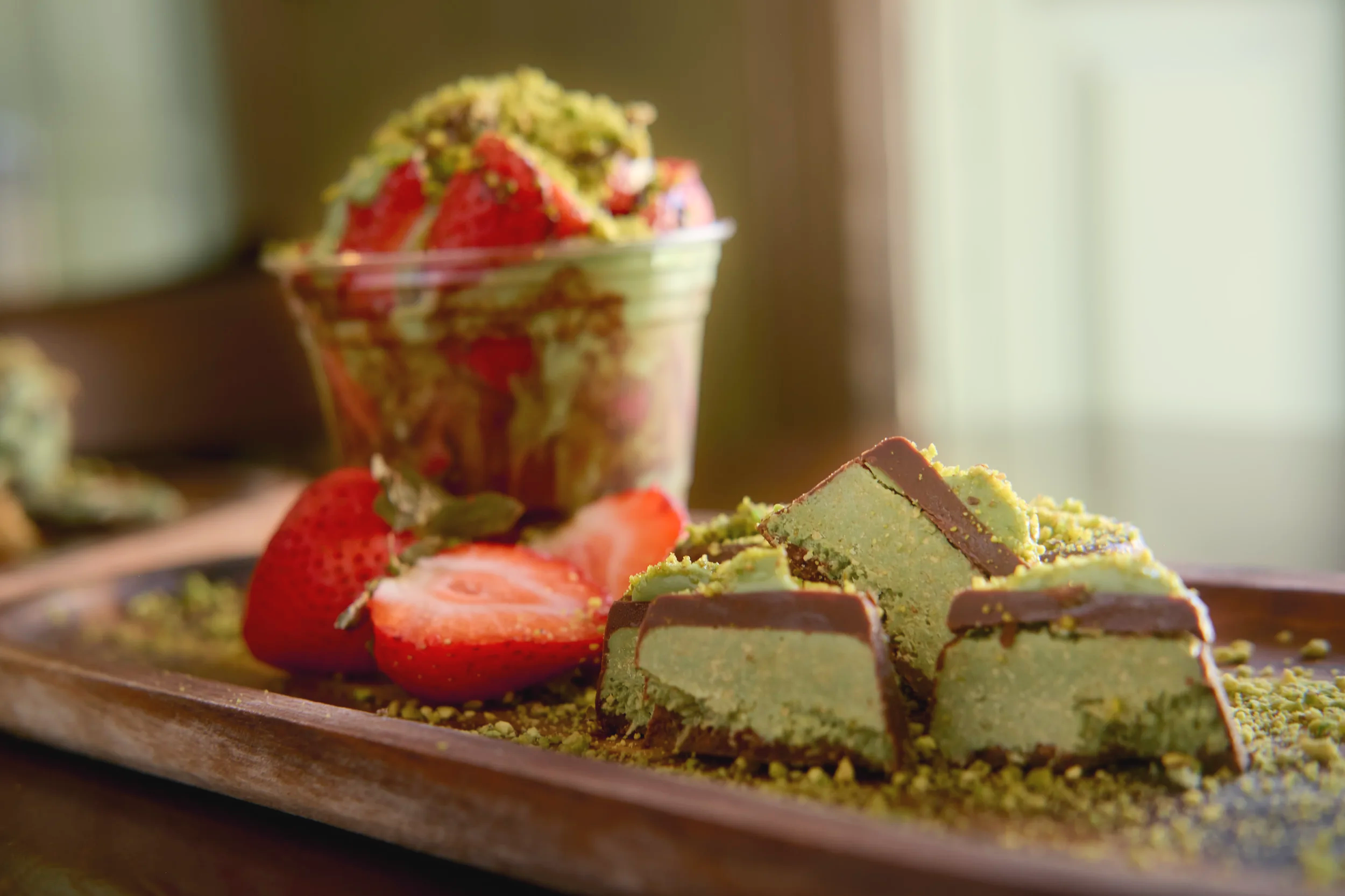 Close-up of a wooden tray with various green and brown chocolate bark pieces, fresh strawberries, and crushed pistachios, with a jar of layered dessert topped with strawberries and pistachios in the background.