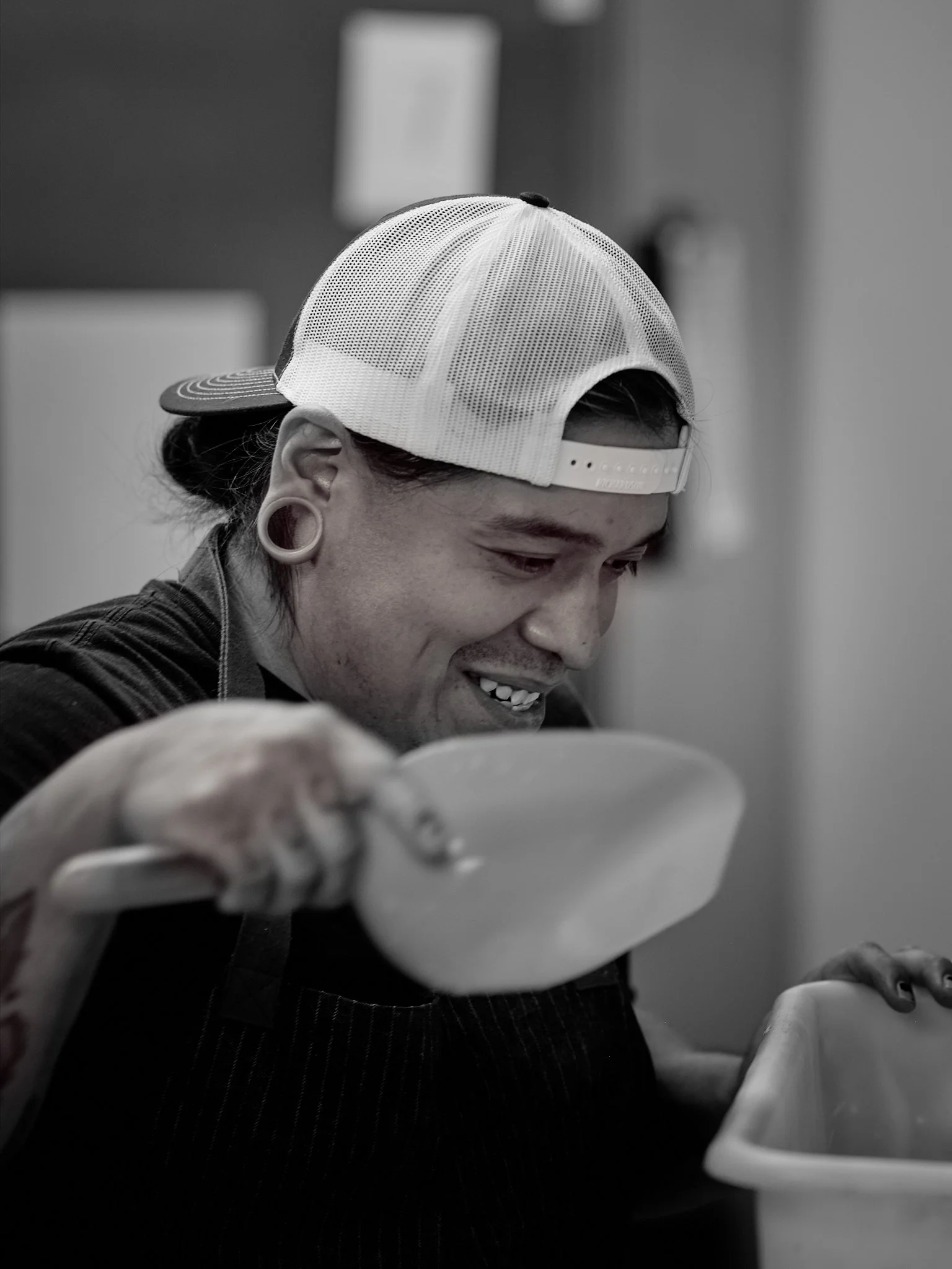 A person with earrings and a baseball cap in a black and white photo, smiling while looking at their phone.