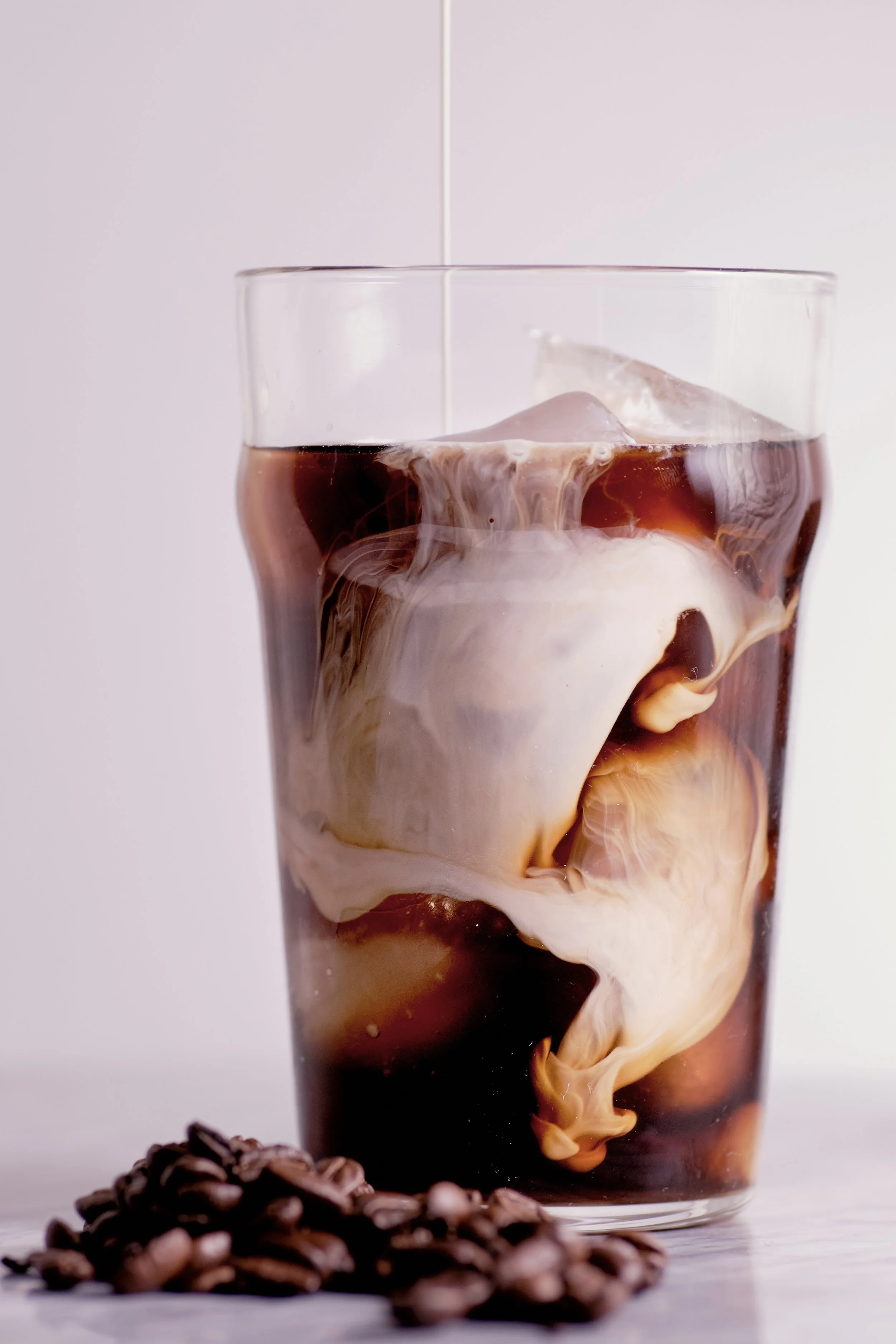 A glass of iced coffee with cream swirling into it, placed on a white surface with coffee beans in the foreground.