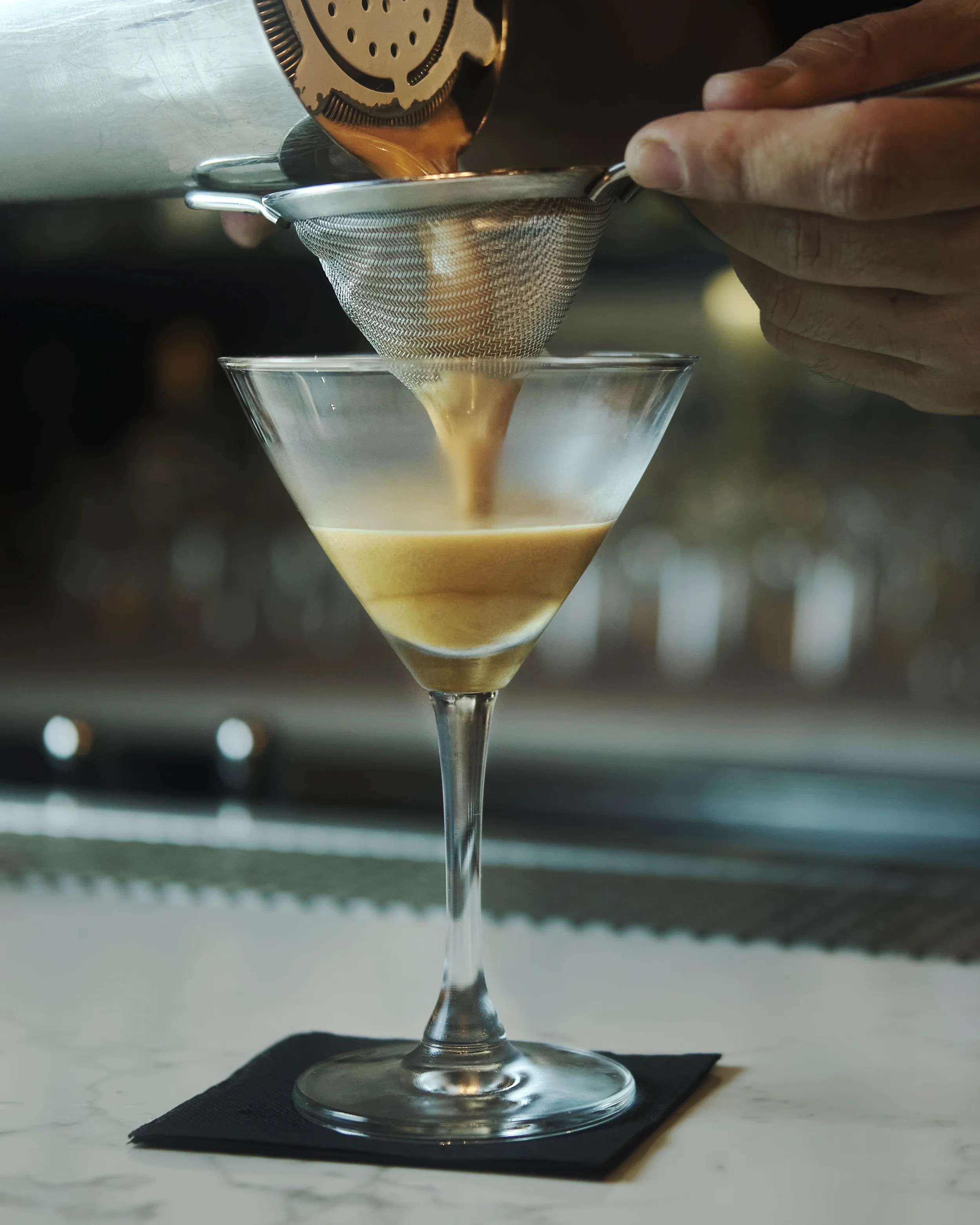 Pouring coffee through a metal sieve into a martini glass on a white surface with a black napkin underneath.