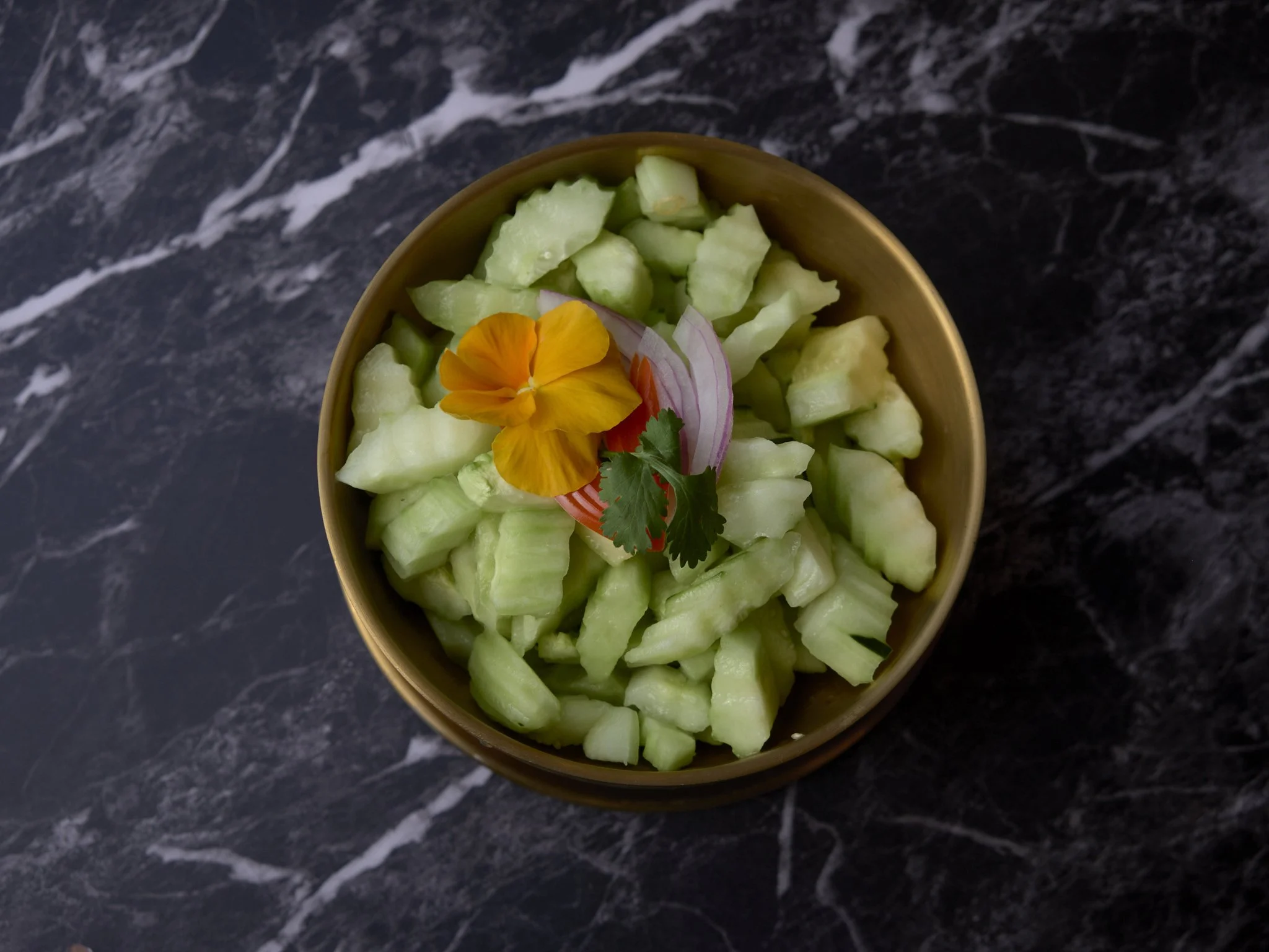 A bowl of chopped cucumber garnished with edible flower, red onion, and cilantro on a black marble surface.