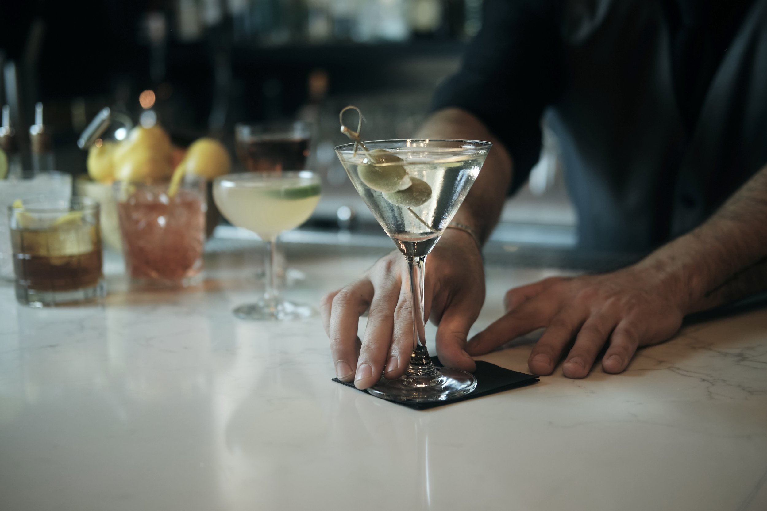 A bartender is preparing a martini glass with olives inside on a bar counter. Various cocktails and garnishes are visible in the background.