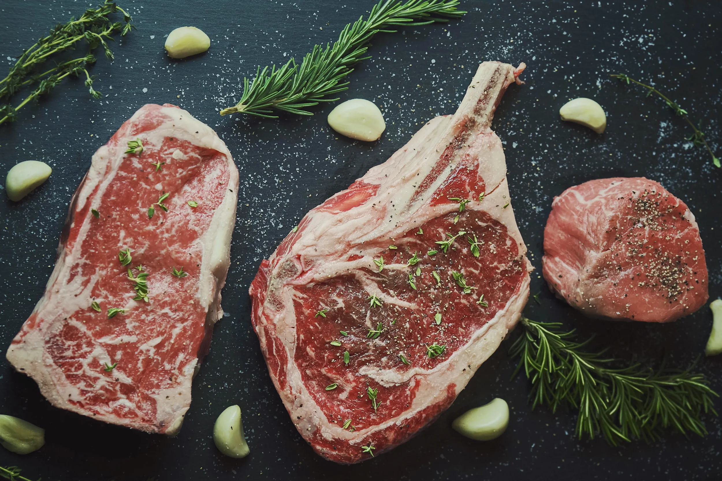 Three uncooked T-bone steaks seasoned with herbs and salt, garnished with fresh rosemary, garlic cloves, and black pepper on a black surface.