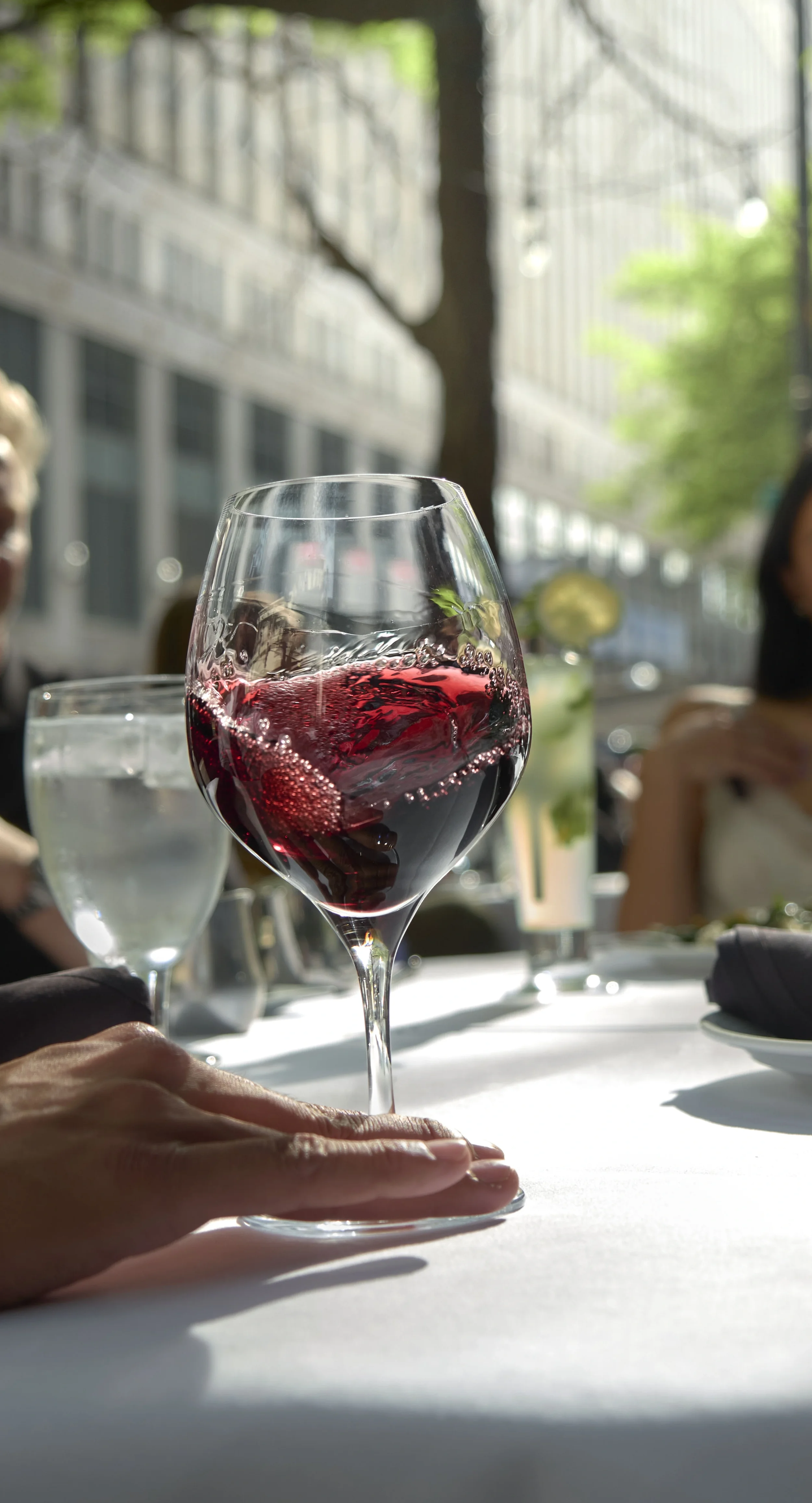 A glass of red wine on a white table at a restaurant, with a background of people dining and large windows letting in natural light.