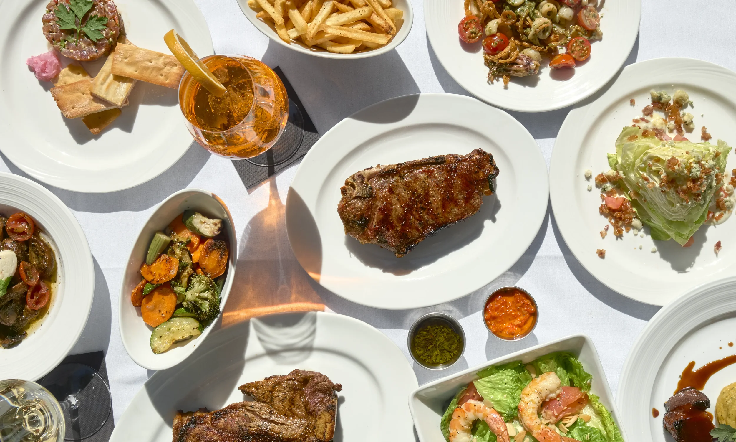 Assorted dishes on white plates including a grilled steak, a salad with shrimp, roasted vegetables, pasta salad, and a glass of orange-colored drink.