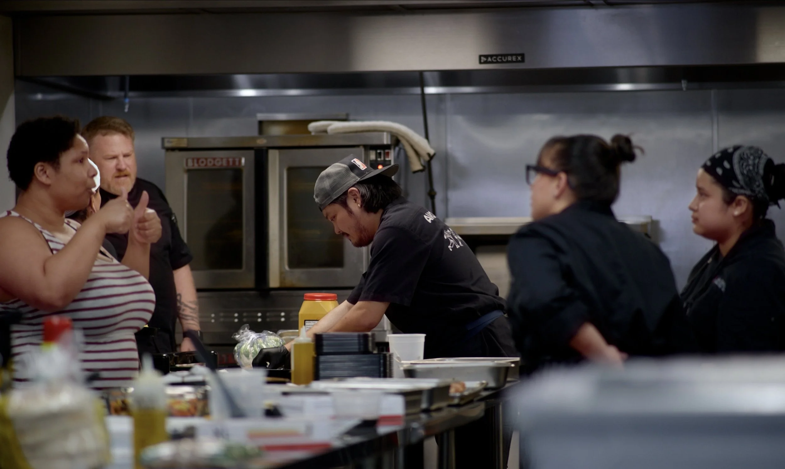 A group of people working in a commercial kitchen, preparing food at a stainless steel counter with various ingredients and condiments. Two women and one man are visible, with the women wearing black uniforms and headscarves, and the man wearing a bl