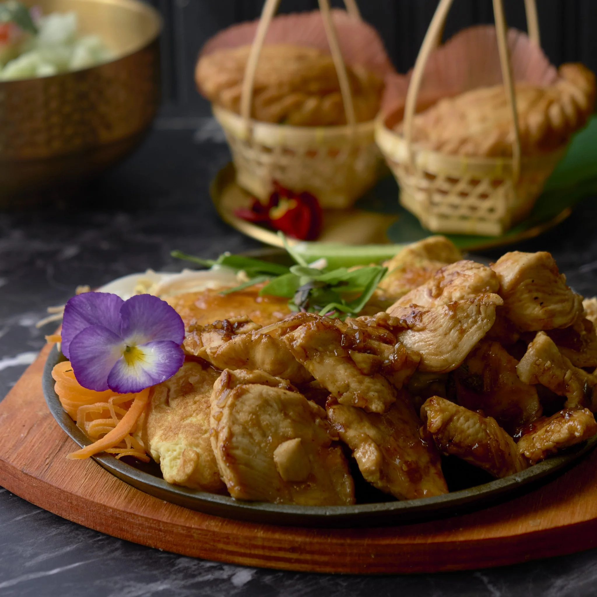 Close-up of a dish with grilled chicken, shredded carrots, microgreens, purple edible flower, and a crispy chip on a black plate with a wooden serving board underneath.