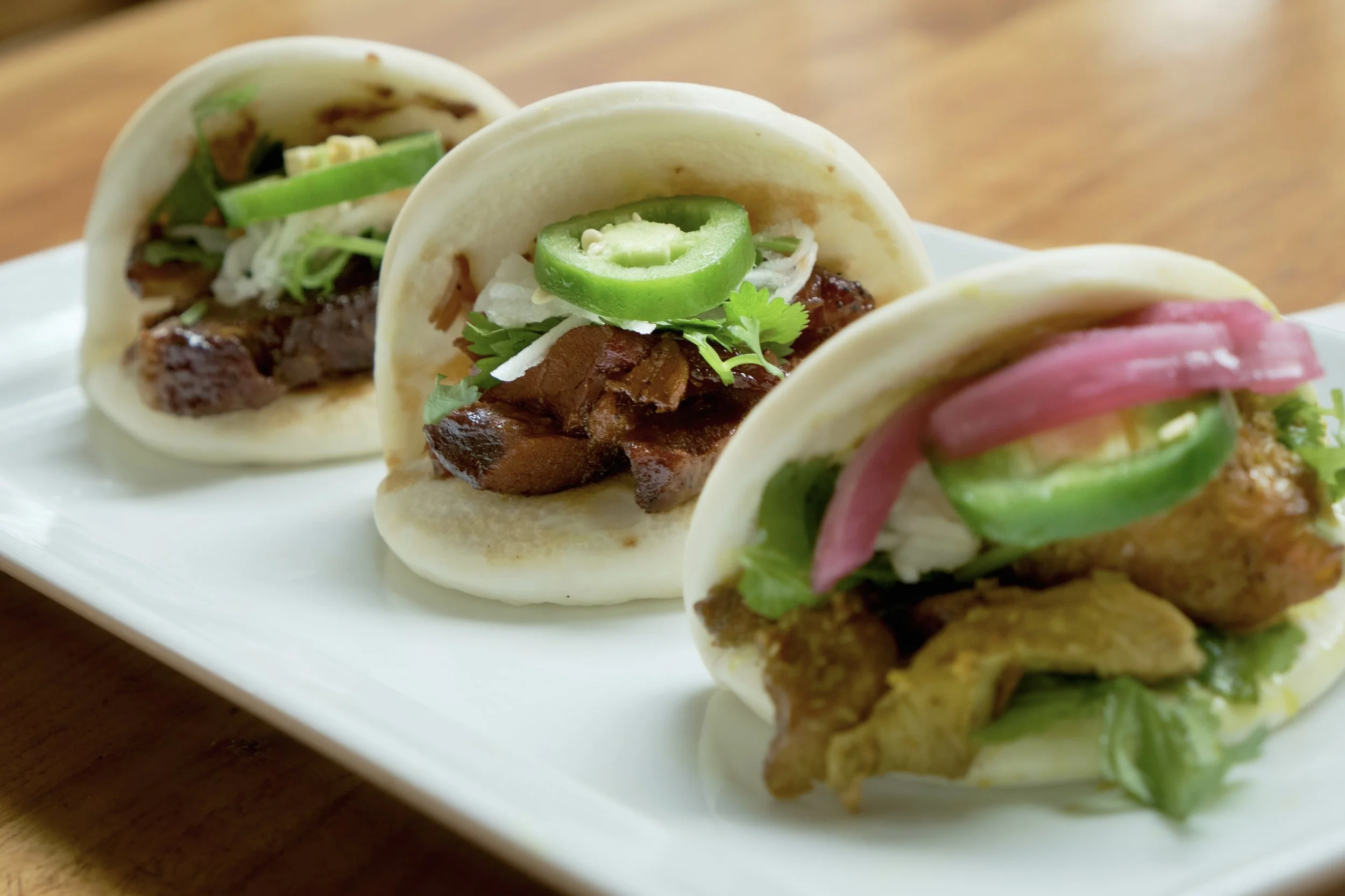 Close-up of three Asian-style tacos with meat, sliced jalapeños, onions, cilantro, and vegetables on a white plate.