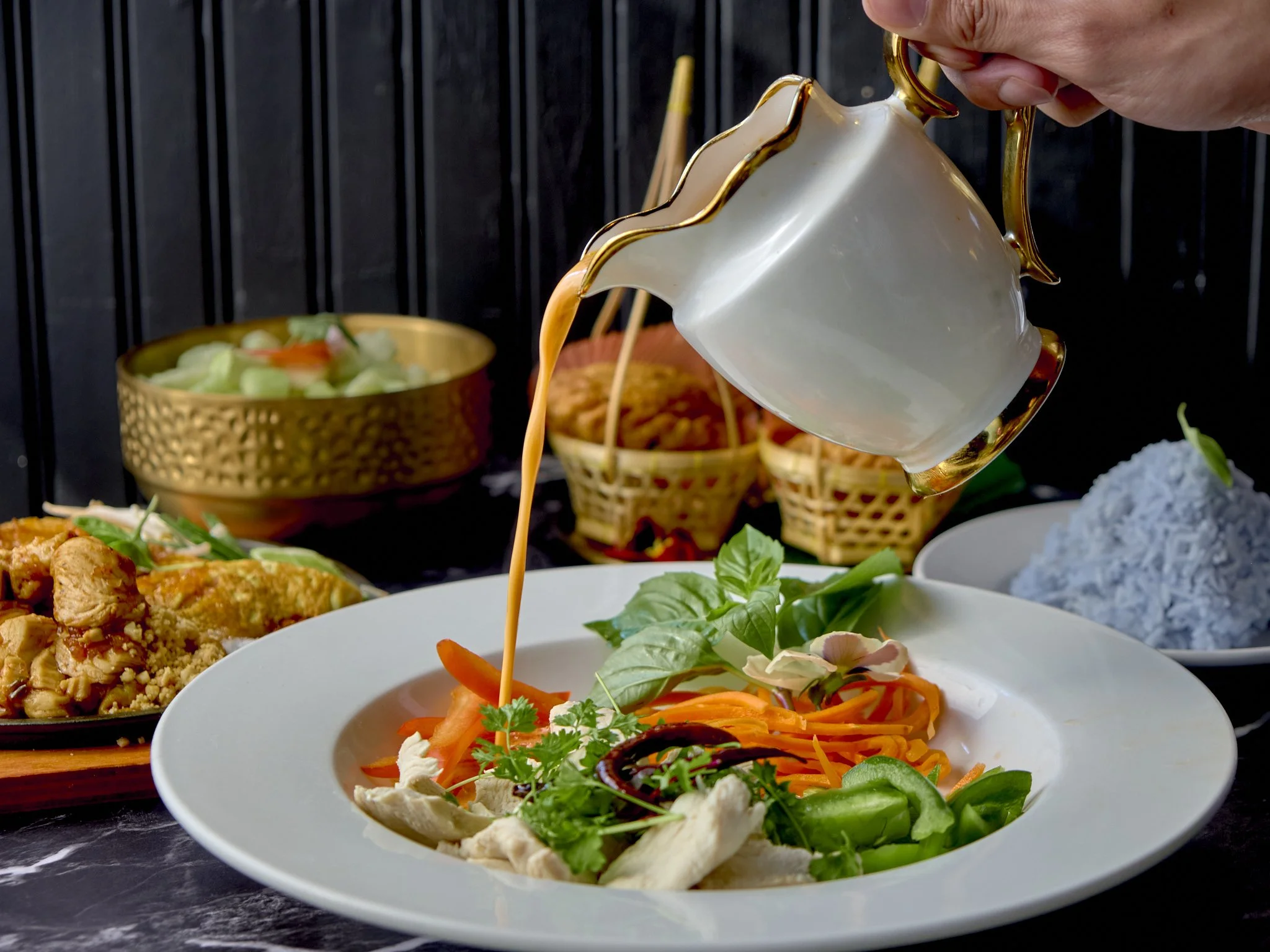 A plate of fresh vegetable salad with carrots, cucumbers, and greens, with a sauce being poured over it from a decorative teapot, with other dishes and bowls in the background.