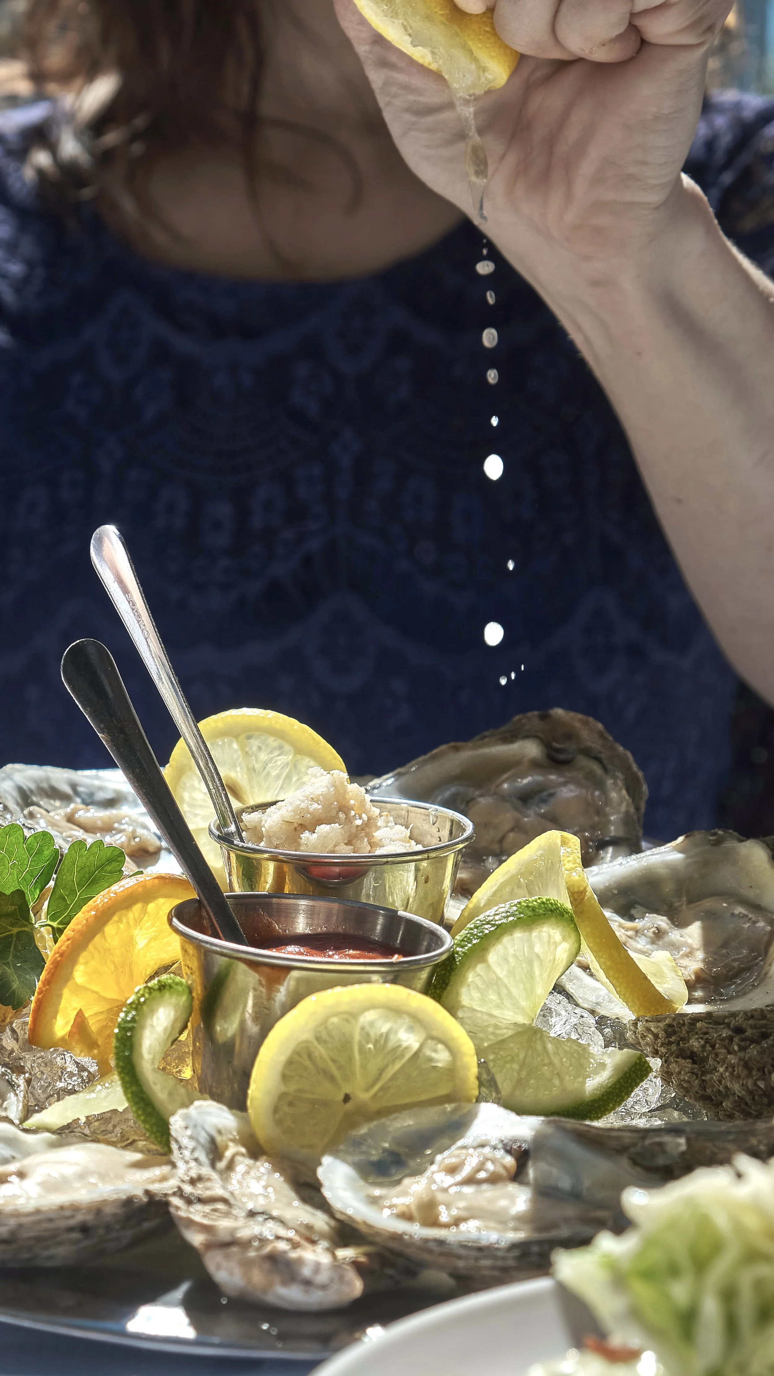 A person sprinkles lemon juice onto oysters on a bed of ice, surrounded by lemon and cucumber slices, with small bowls of condiments nearby.