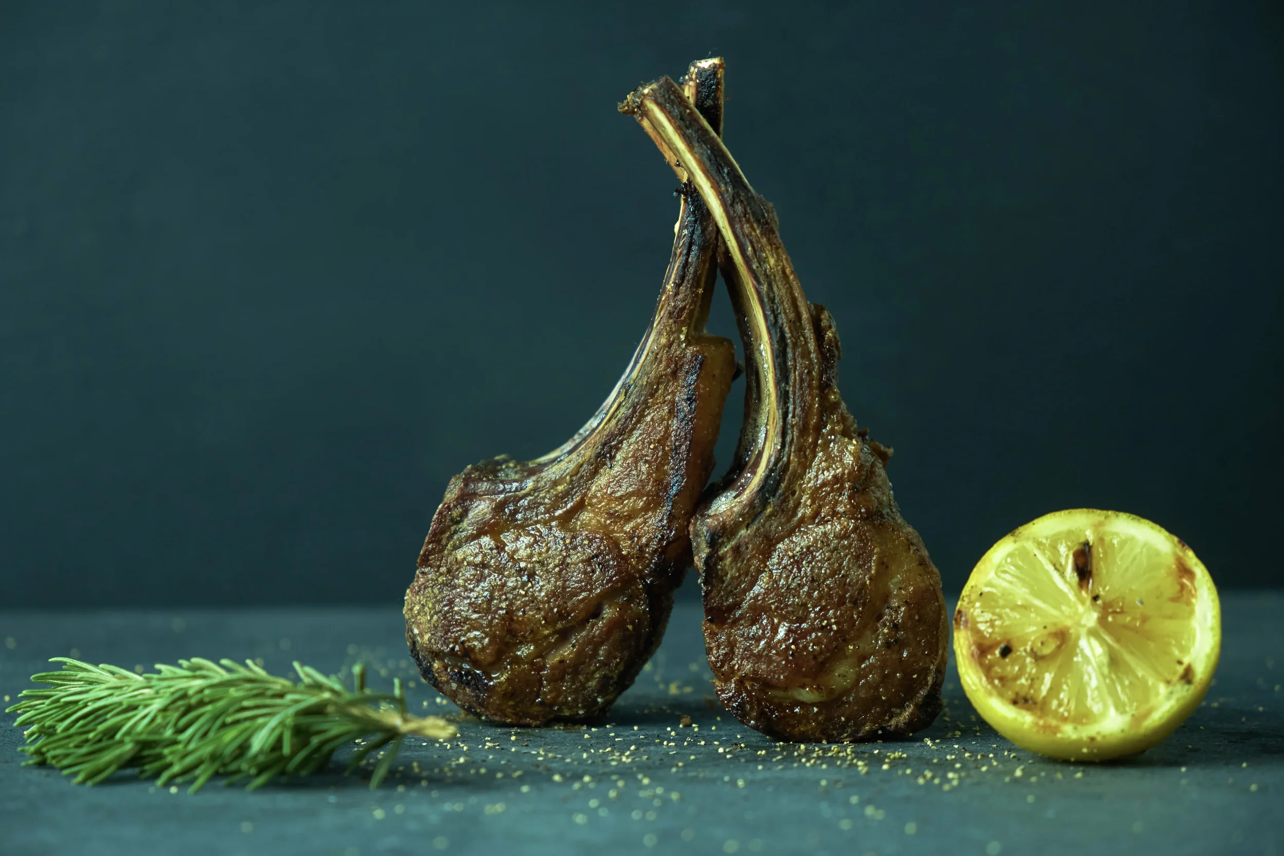 A close-up of two cooked lamb chops stacked vertically, garnished with lemon slices, placed on a dark surface with sprigs of rosemary and scattered seasonings around.