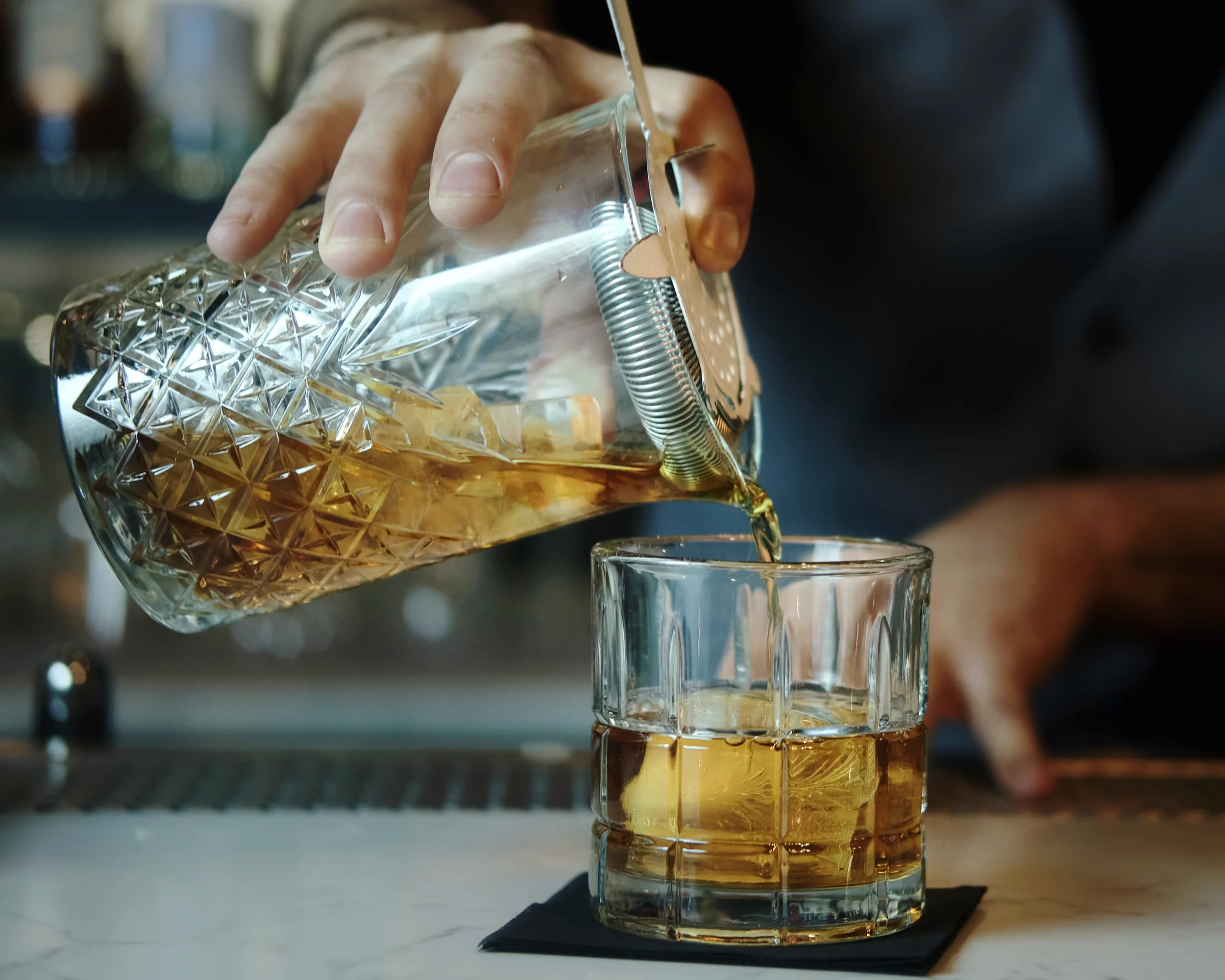 A close-up of a hand holding a crystal glass with a gold-colored liquid, pouring it into a short glass in a dimly-lit setting.