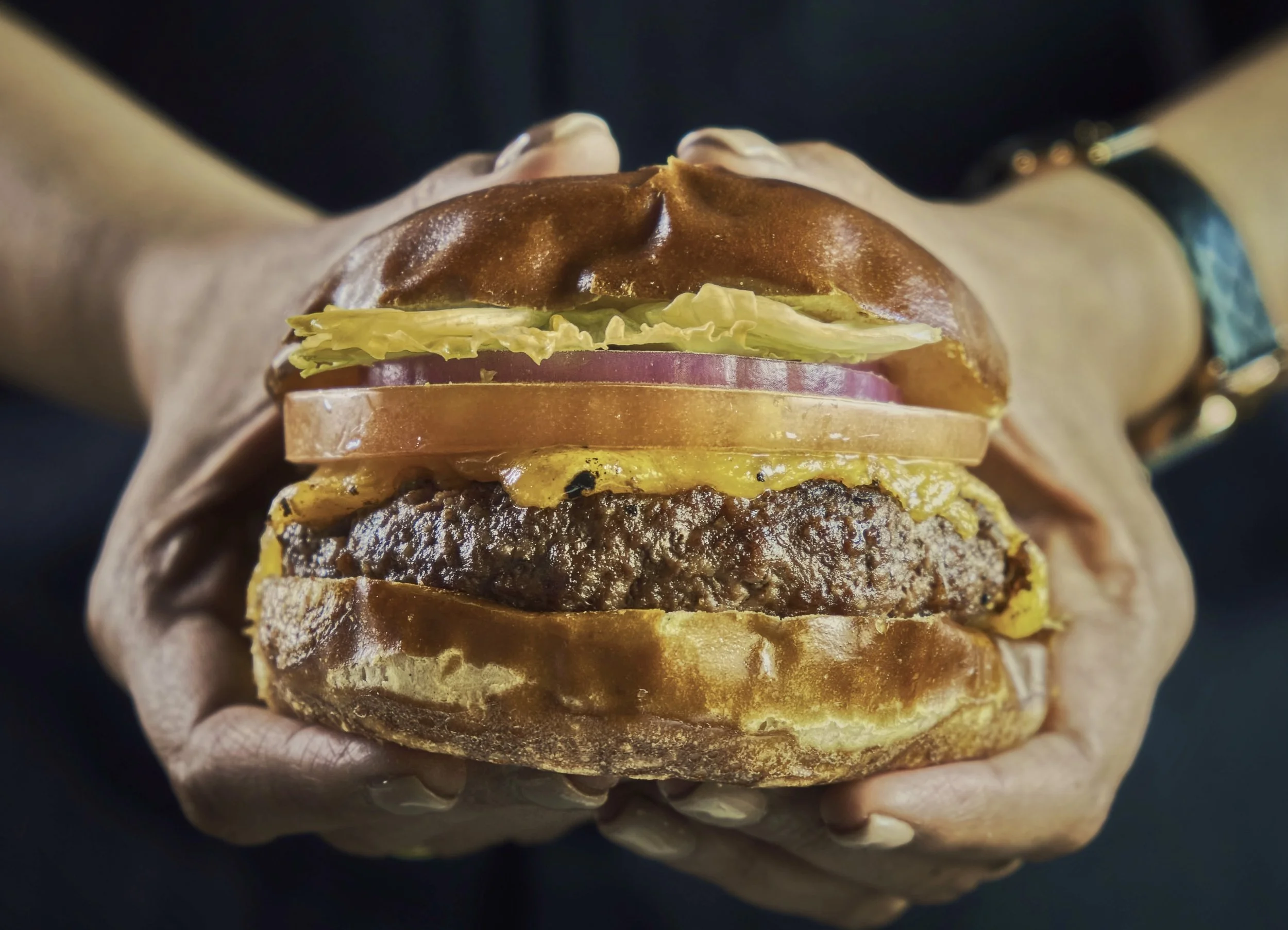 Close-up of a large, layered cheeseburger held in a person's hand. The burger includes lettuce, tomato, onion, a beef patty, cheese, and a toasted bun.